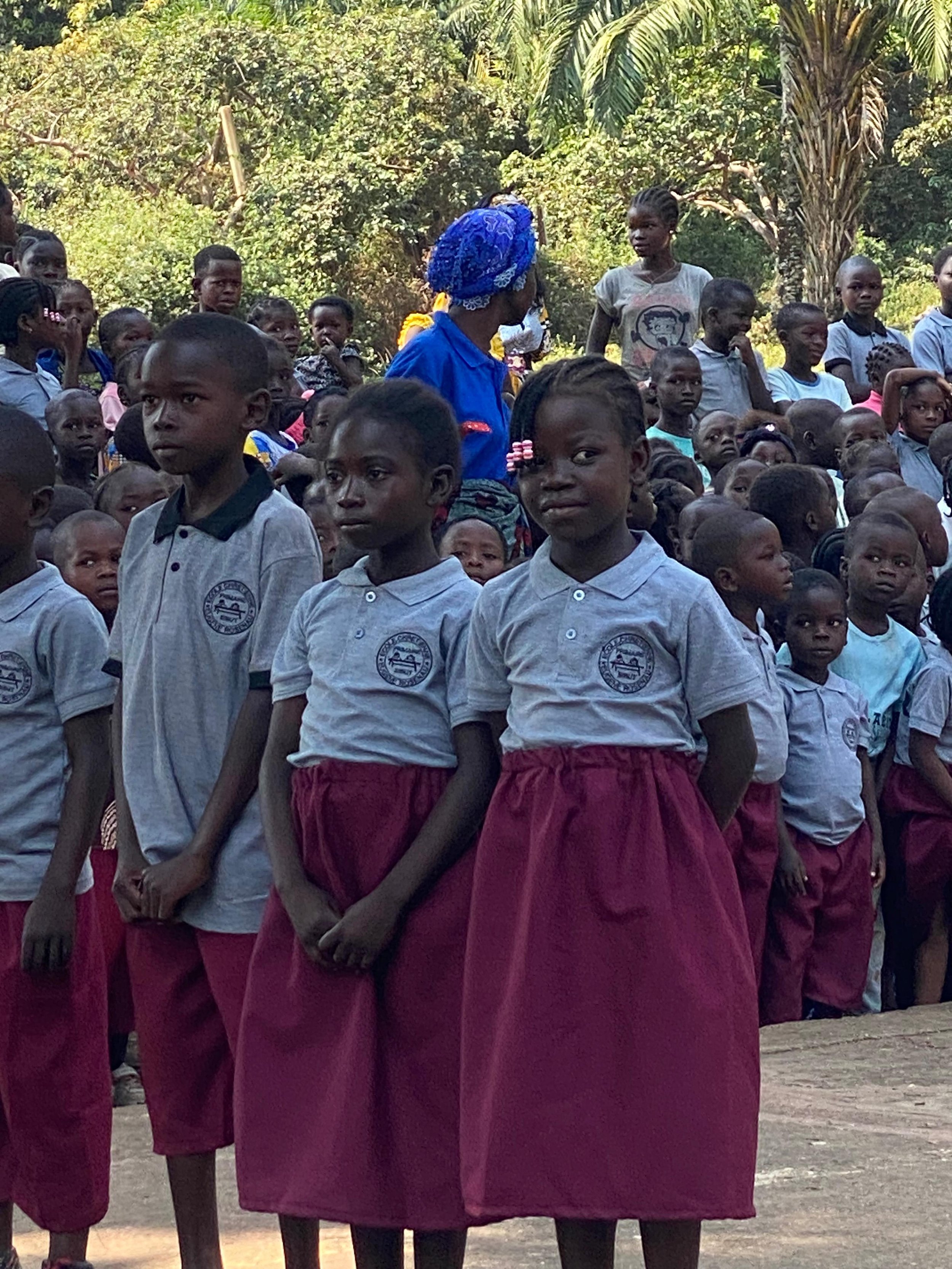 Group of children in school uniforms standing outdoors, some children are looking at the camera and others looking away, with trees in the background.