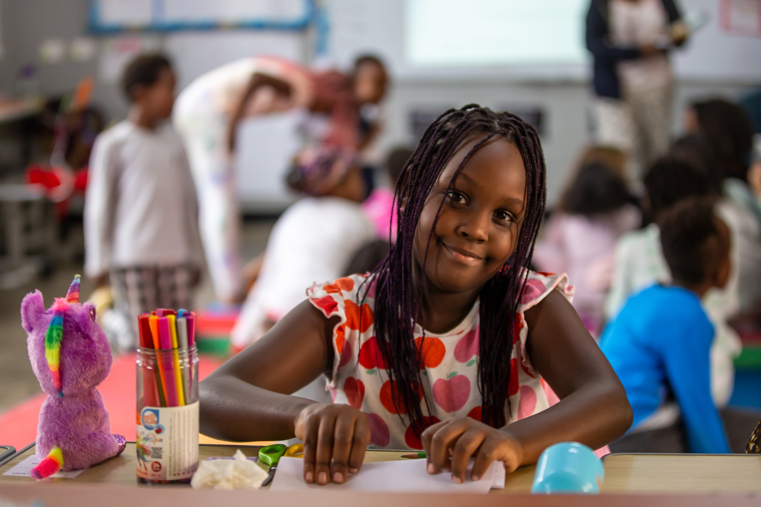 A young girl with dressed in a polka-dot shirt, smiling at the camera, sitting at a table with coloring supplies in a classroom full of children in the background.