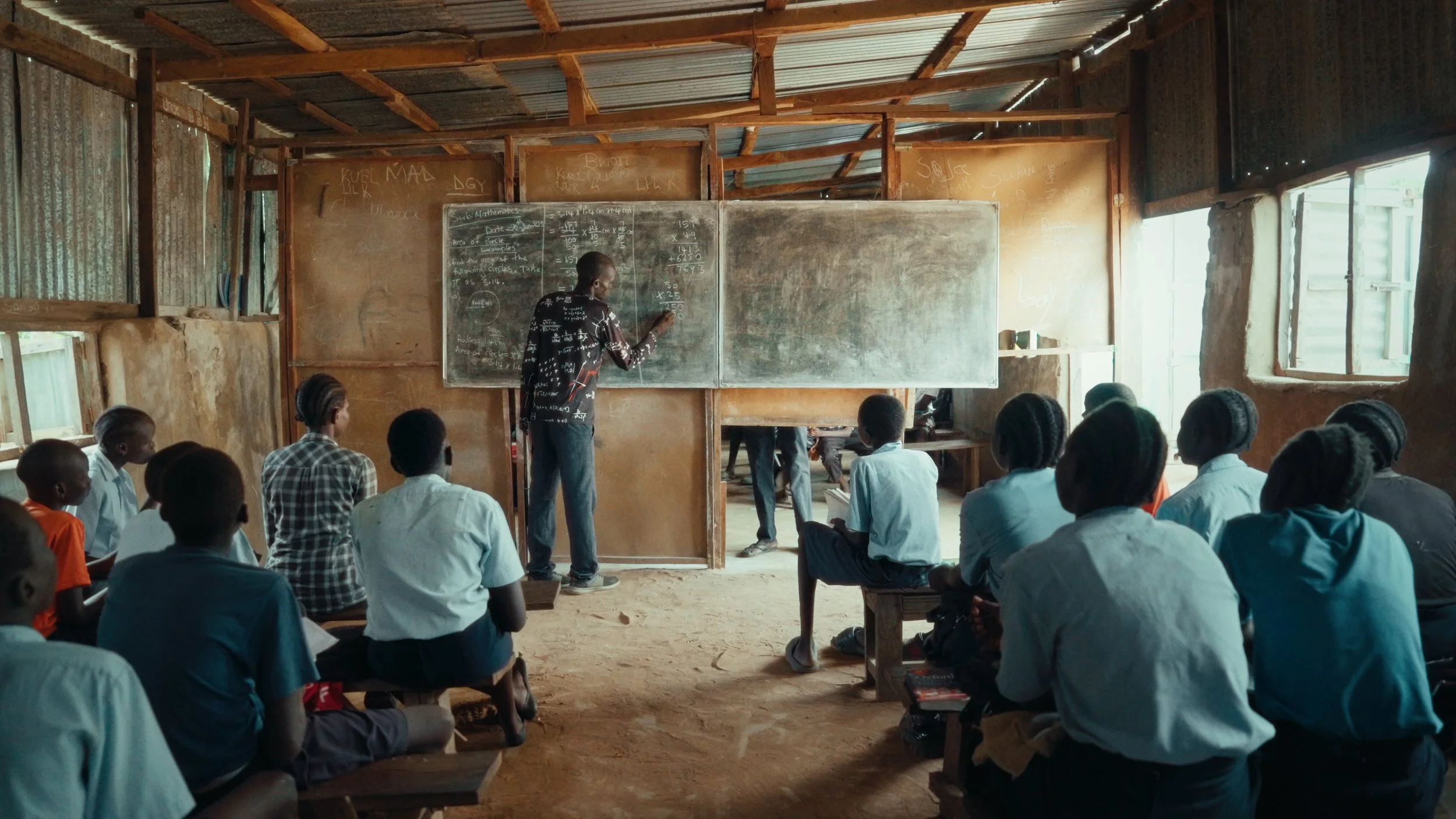 A classroom with students sitting on benches and a teacher writing on a chalkboard inside a building with wooden walls and tin roof.