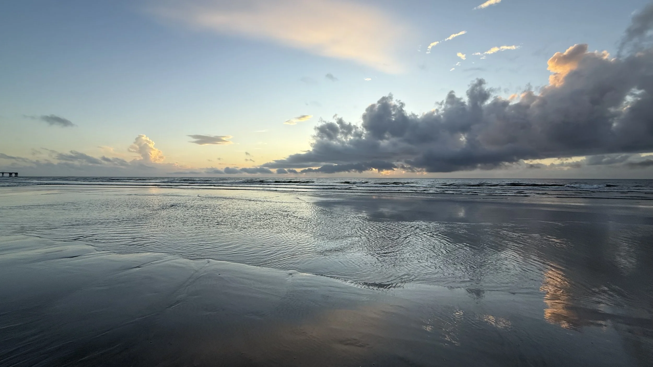A serene beach scene at sunset with the sky partly cloudy, reflecting on the wet sand and gentle ocean waves.