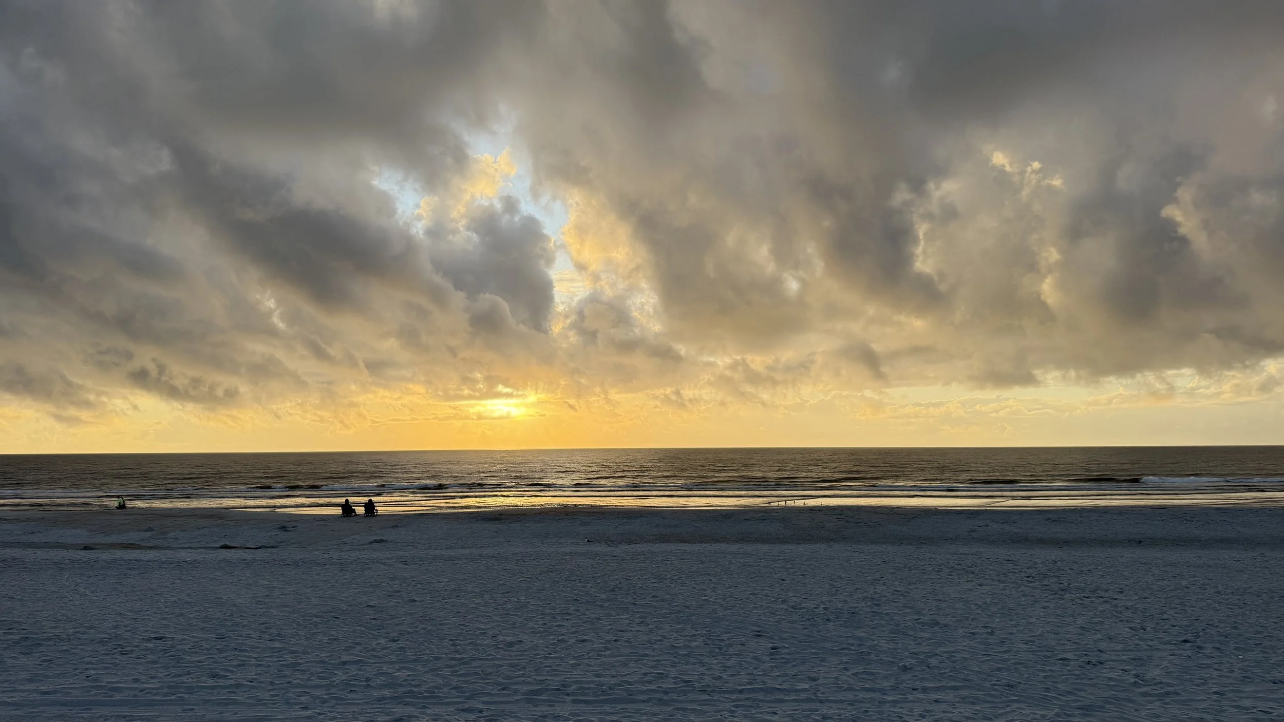 A beach scene during sunset with dark clouds overhead, the sun partially visible near the horizon, and a few people sitting on the sand near the water.