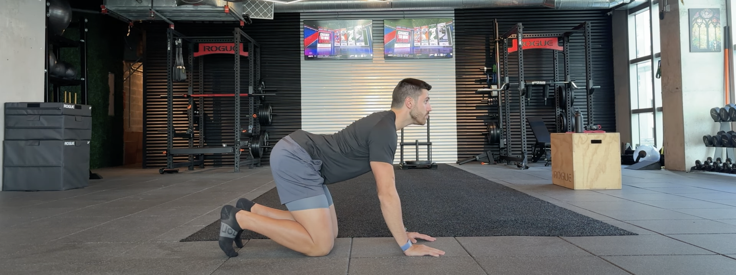 A man performing a tabletop exercise in a gym, on his hands and knees on a black exercise mat, with gym equipment and two television screens in the background.