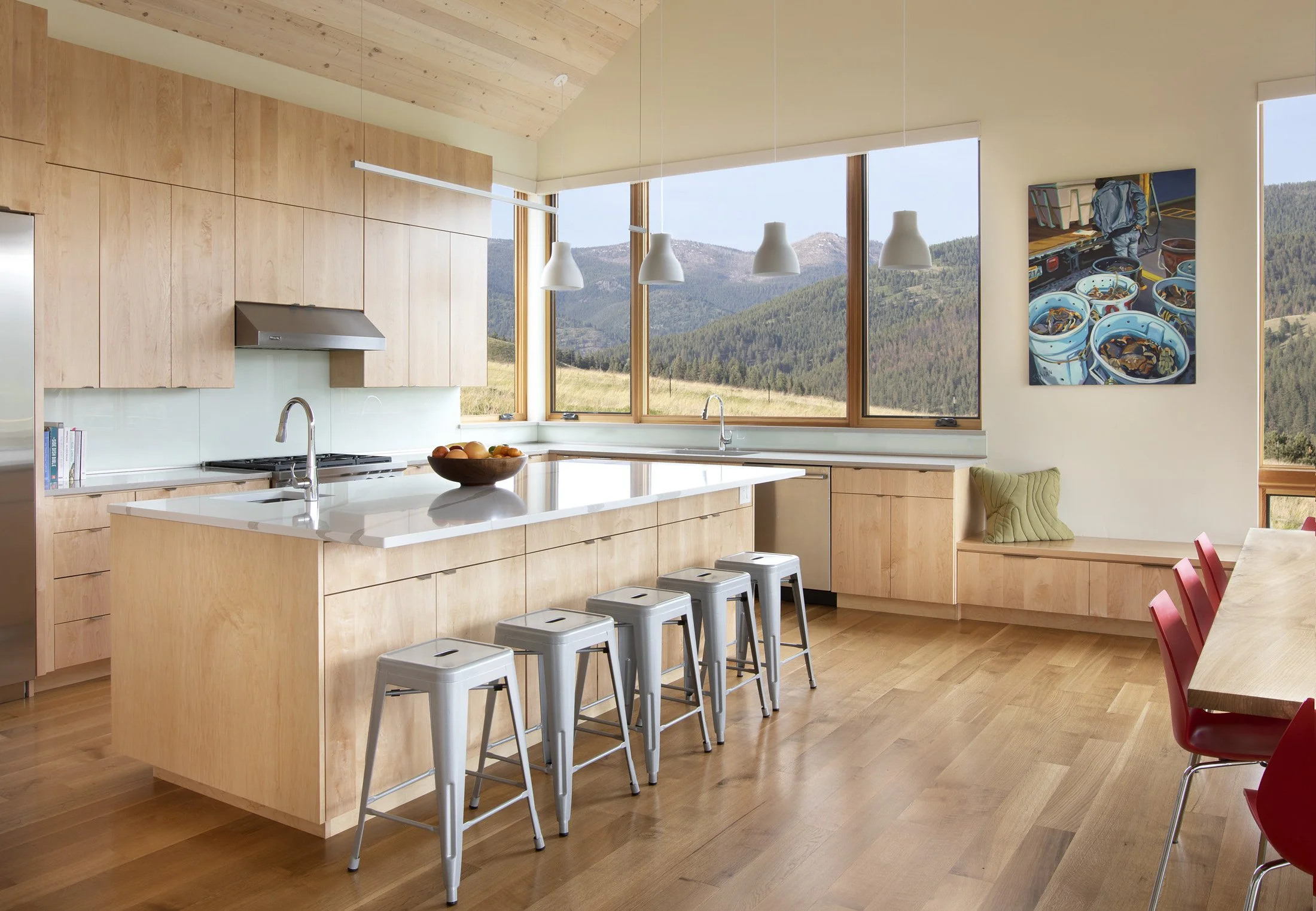 A modern kitchen with light wooden cabinets and a white island with stools, large windows with mountain views, a chandelier, and a dining area with red chairs. Custom Home built in Montana by Edgell Building.