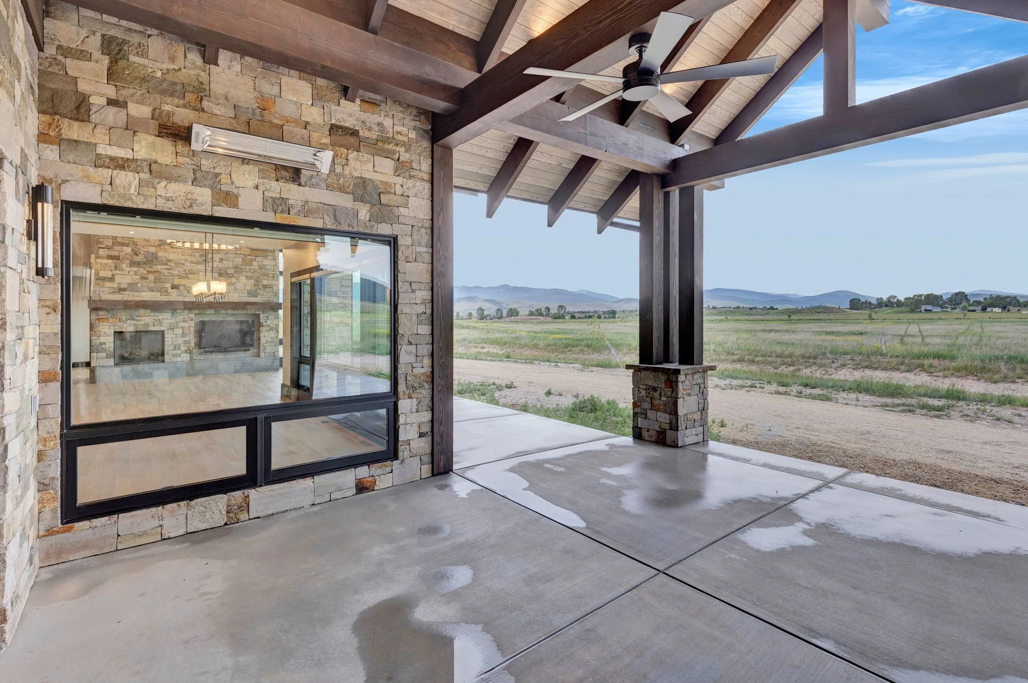 Covered patio with stone walls, wooden beams, ceiling fan, and a view of fields and mountains in the distance, with a wet concrete floor. Custom Home built in Montana by Edgell Building.