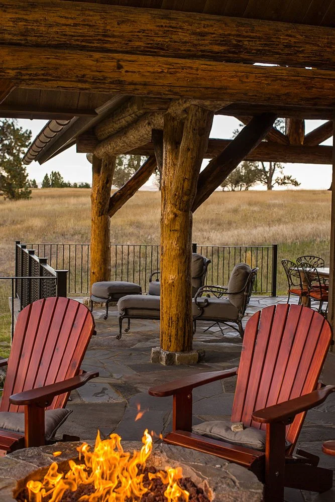 A rustic outdoor patio with red Adirondack chairs around a fire pit, wooden beams supporting a roof, cushioned lounge chairs, and a view of open fields with trees in the distance. Custom Home built in Montana by Edgell Building.