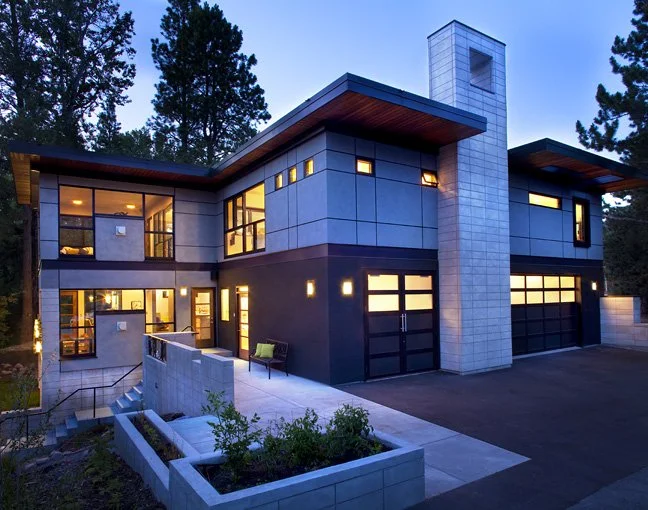 Modern two-story house with large windows, a garage, and a chimney, surrounded by trees at dusk. Custom Home built in Montana by Edgell Building.