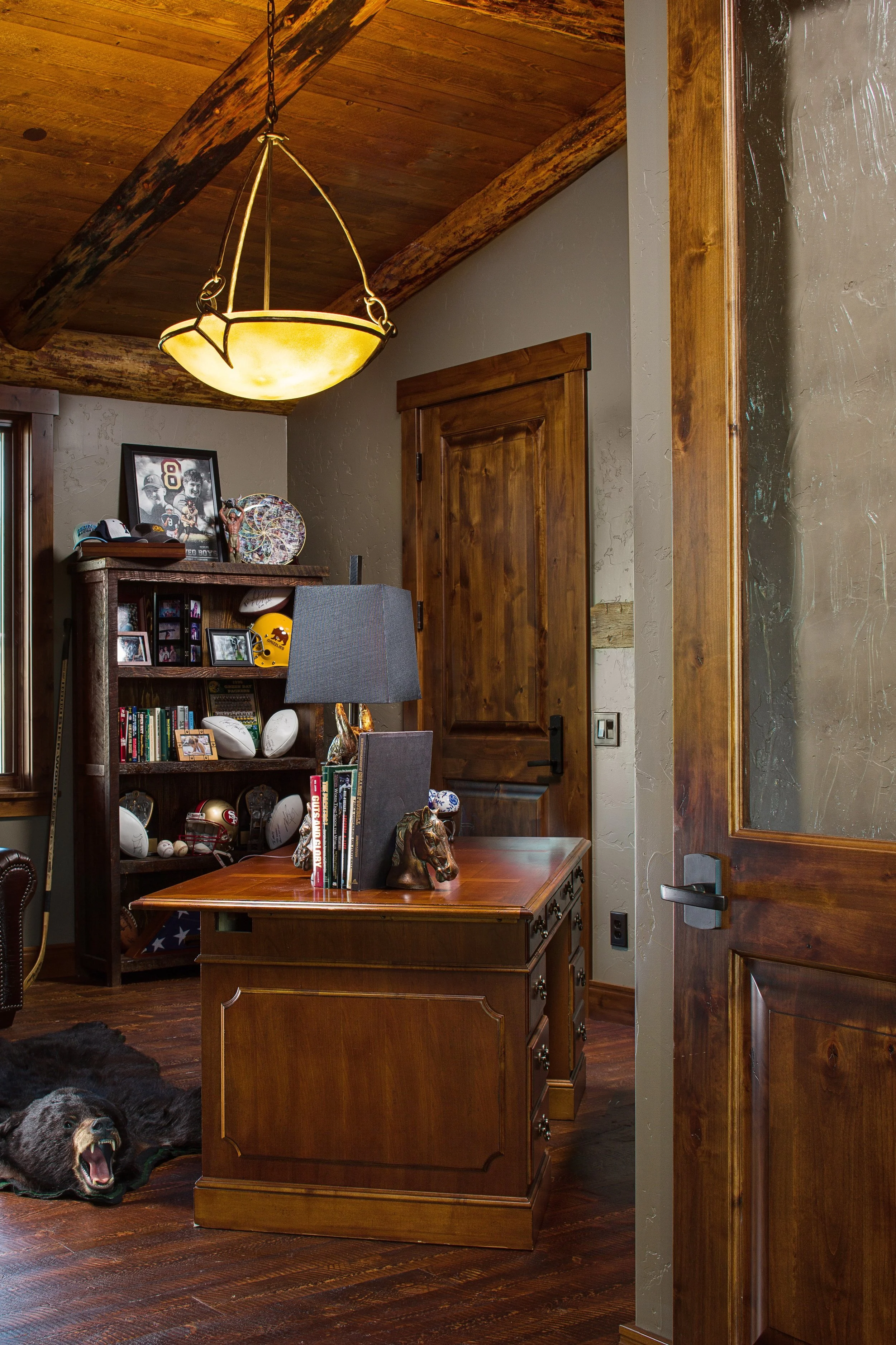 A cozy office with wooden furniture, sports memorabilia, a bookshelf filled with books and footballs, a modern lamp, and a large wooden door. The floor and ceiling are also made of wood, creating a warm atmosphere. Custom Home built in Montana by Edg