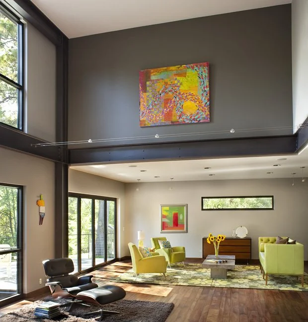 A modern living room with large windows, colorful abstract artwork on a gray wall, green chairs, a wooden sideboard, and decorative vases and art pieces. Custom Home built in Montana by Edgell Building.