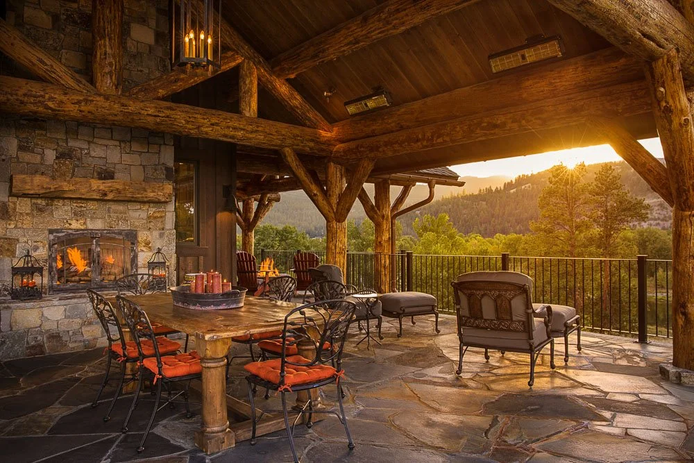A cozy outdoor patio with a stone fireplace, wooden beams, andmountain view, furnished with a rustic dining table, chairs with orange cushions, and lounge chairs, illuminated by warm lighting and a sunset. Custom Home built in Montana by Edgell Build