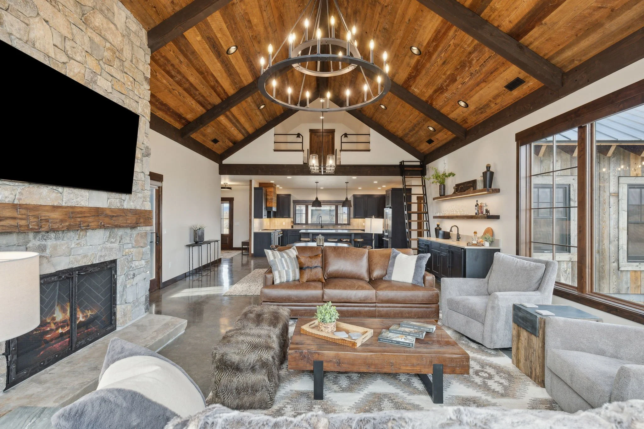Living room with vaulted wood ceiling, stone fireplace, leather sofa, gray armchairs, large windows, and open kitchen in the background. Custom Home built in Montana by Edgell Building.