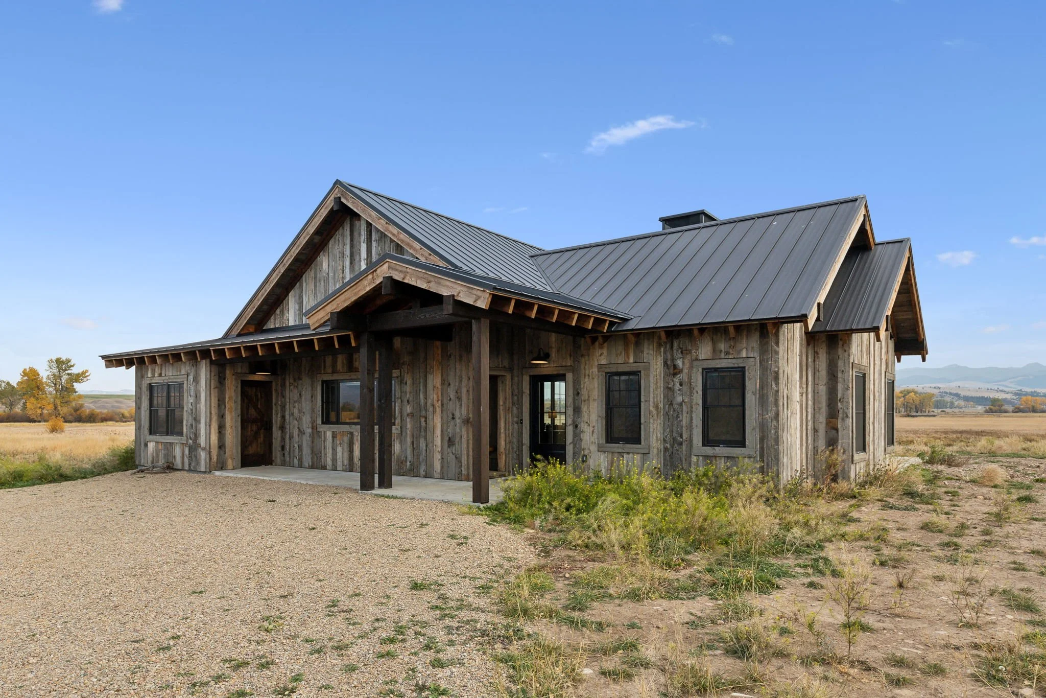 A rustic, weathered wooden house with a metal roof, set in a rural landscape under a blue sky, with sparse vegetation around a gravel driveway. Custom Home built in Montana by Edgell Building.