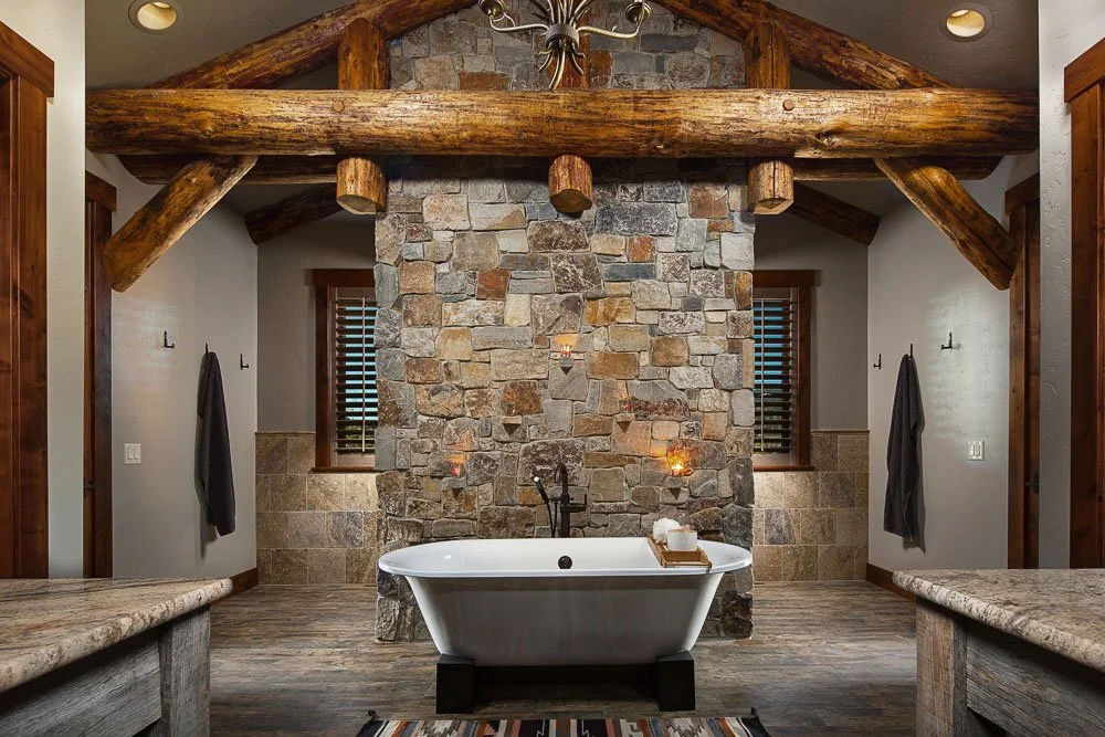 A rustic bathroom with a freestanding bathtub in front of a stone accent wall, wooden beams on the ceiling, and two small windows with shutters. Custom Home built in Montana by Edgell Building.