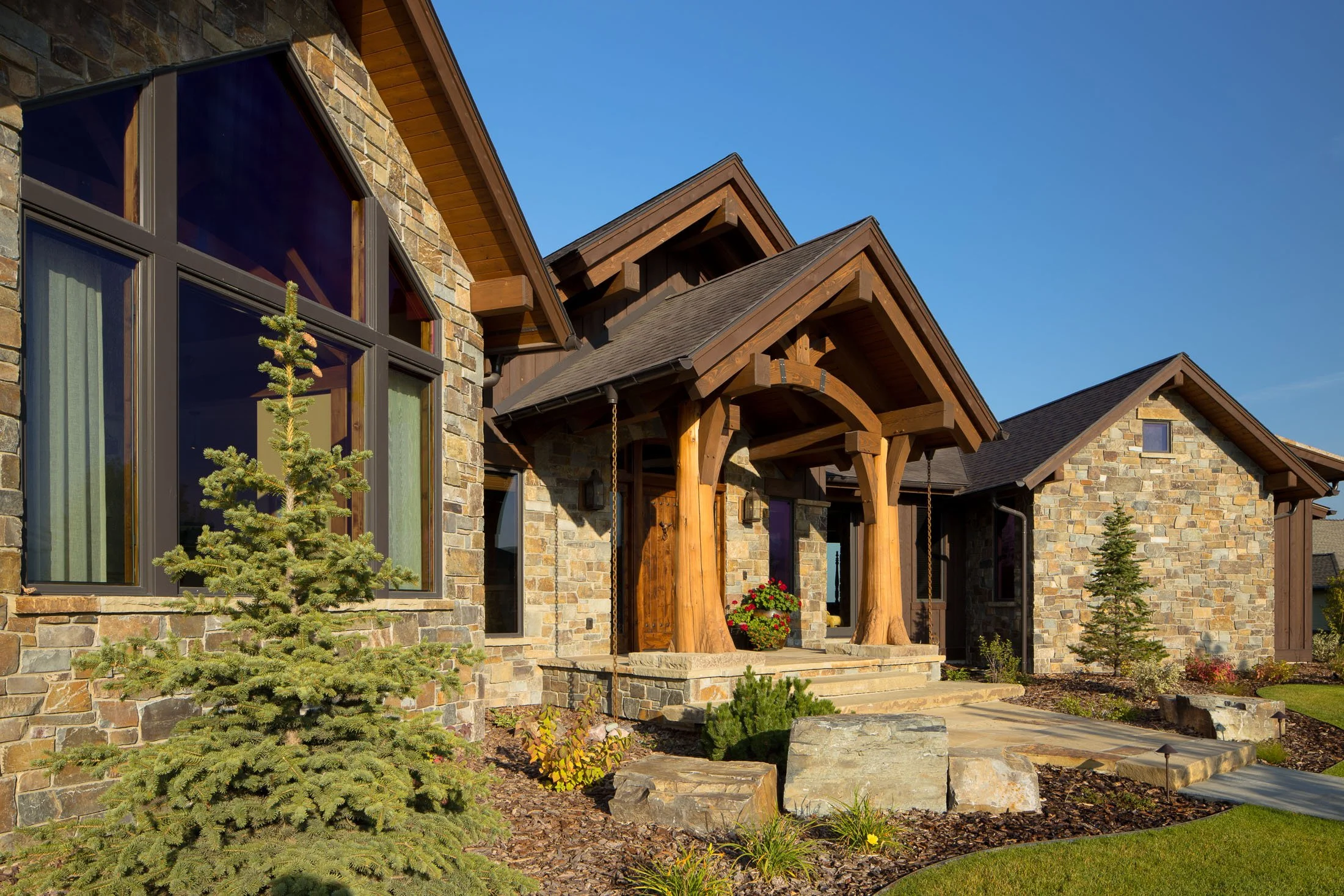 Front exterior view of a house with stone and wood siding, large reflective windows, a covered porch with wooden posts, steps leading to the entrance, and landscaped yard with small trees and plants. Custom Home built in Montana by Edgell Building.