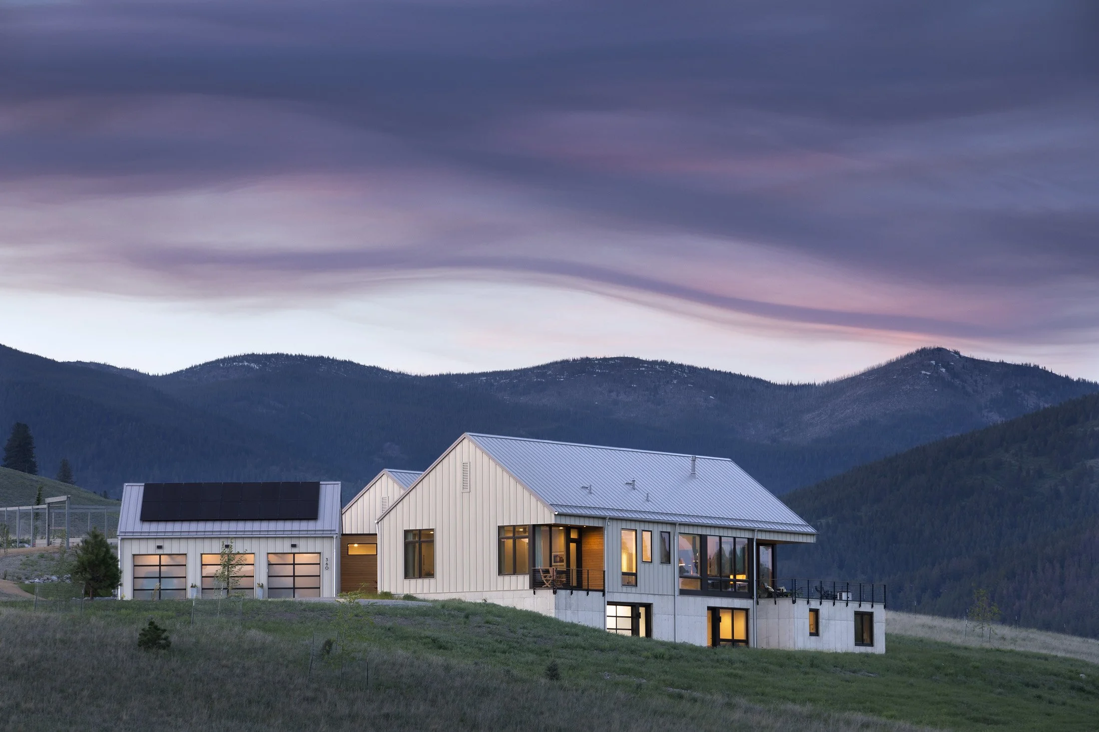 Modern house with white siding, large windows, and a metal roof, situated on a grassy hill with mountains in the background and a colorful sky during dusk. Custom Home built in Montana by Edgell Building.