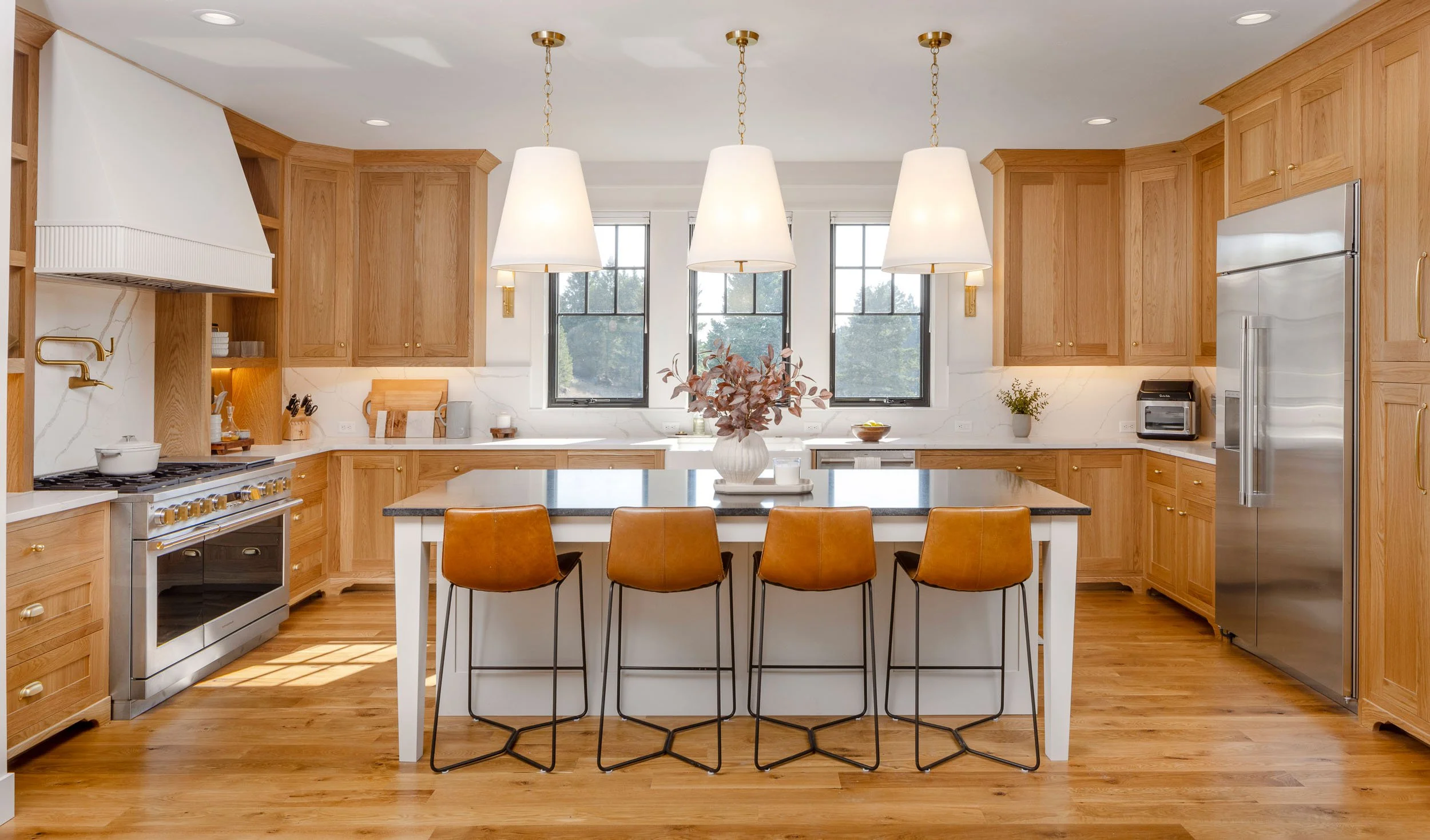Modern kitchen with wooden cabinets, stainless steel appliances, a large island with four brown leather chairs, white countertops, three white pendant lights, and a vase with pinkish leaves on the island. Custom Home in Montana by Edgell Building.