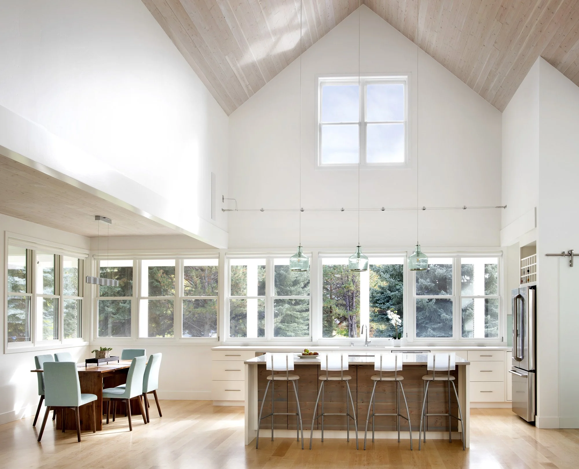 Bright and spacious kitchen with large windows, wooden ceiling, central island with chairs, and a dining area with a round wooden table and six chairs. Custom Home built in Montana by Edgell Building.