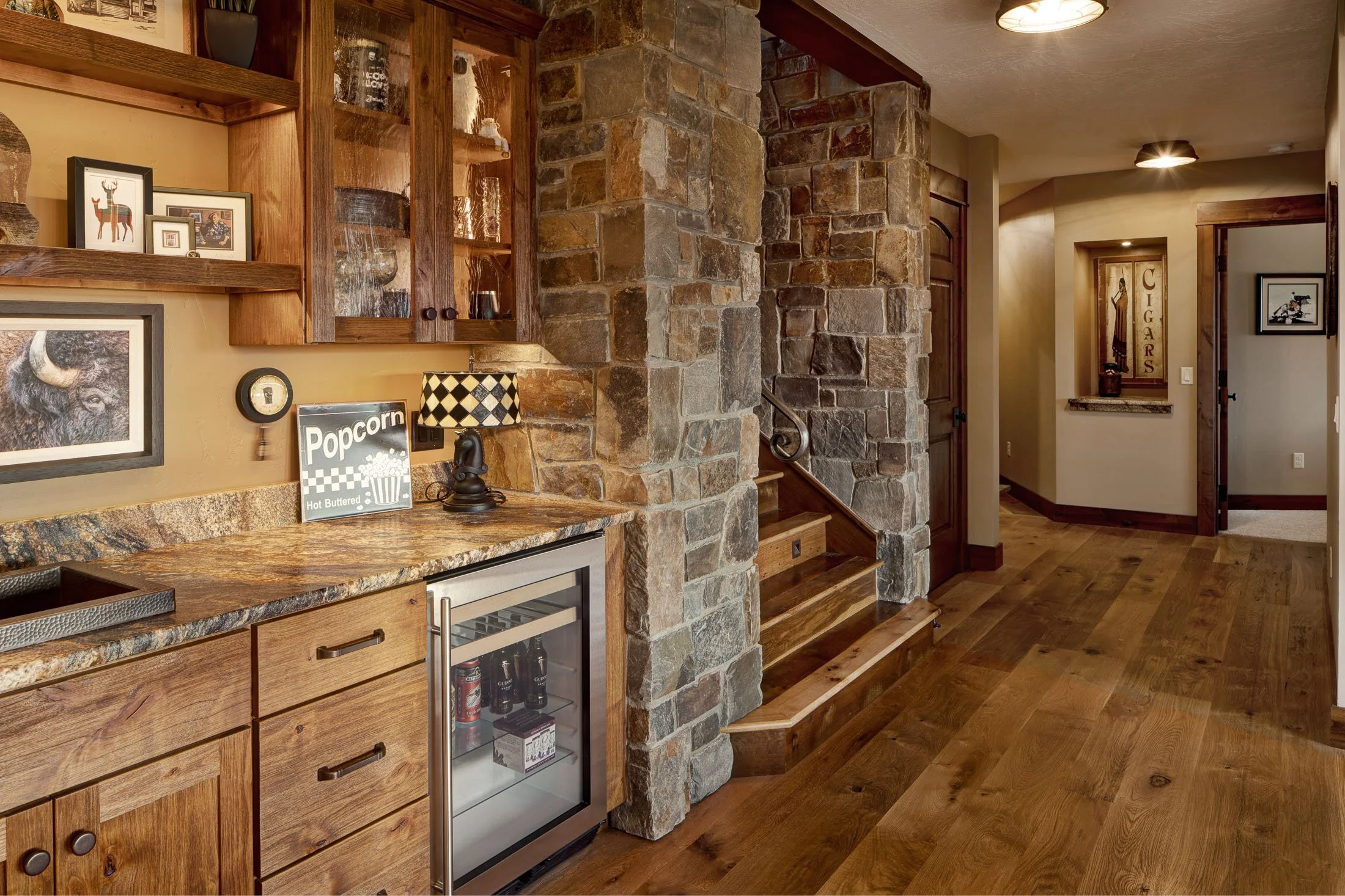 Interior of a rustic home with wooden floors and walls, a stone staircase, a small wet bar with a granite countertop, and decorative artwork including framed pictures and signs. Custom Home built in Montana by Edgell Building.