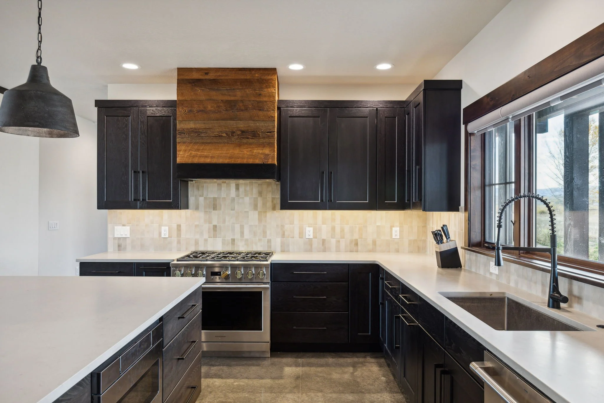Modern kitchen with black cabinets, beige backsplash, stainless steel stove, and a large window with a black faucet. Custom Home built in Montana by Edgell Building.