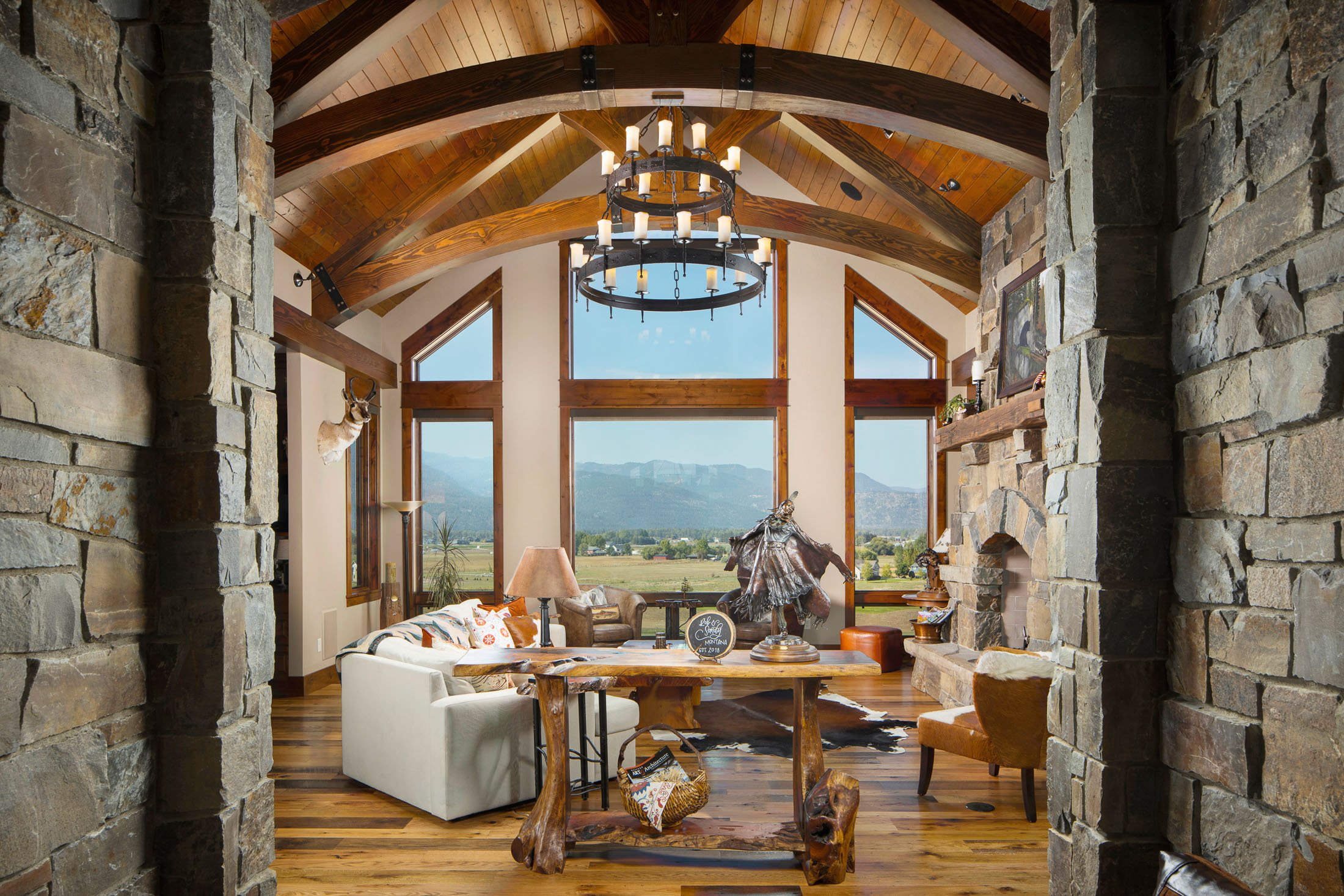A spacious living room with high vaulted wooden ceilings, large windows, a stone fireplace, and rustic decor including a deer head mount, a wooden table, a white sofa, and a chandelier. Custom Home built in Montana by Edgell Building.