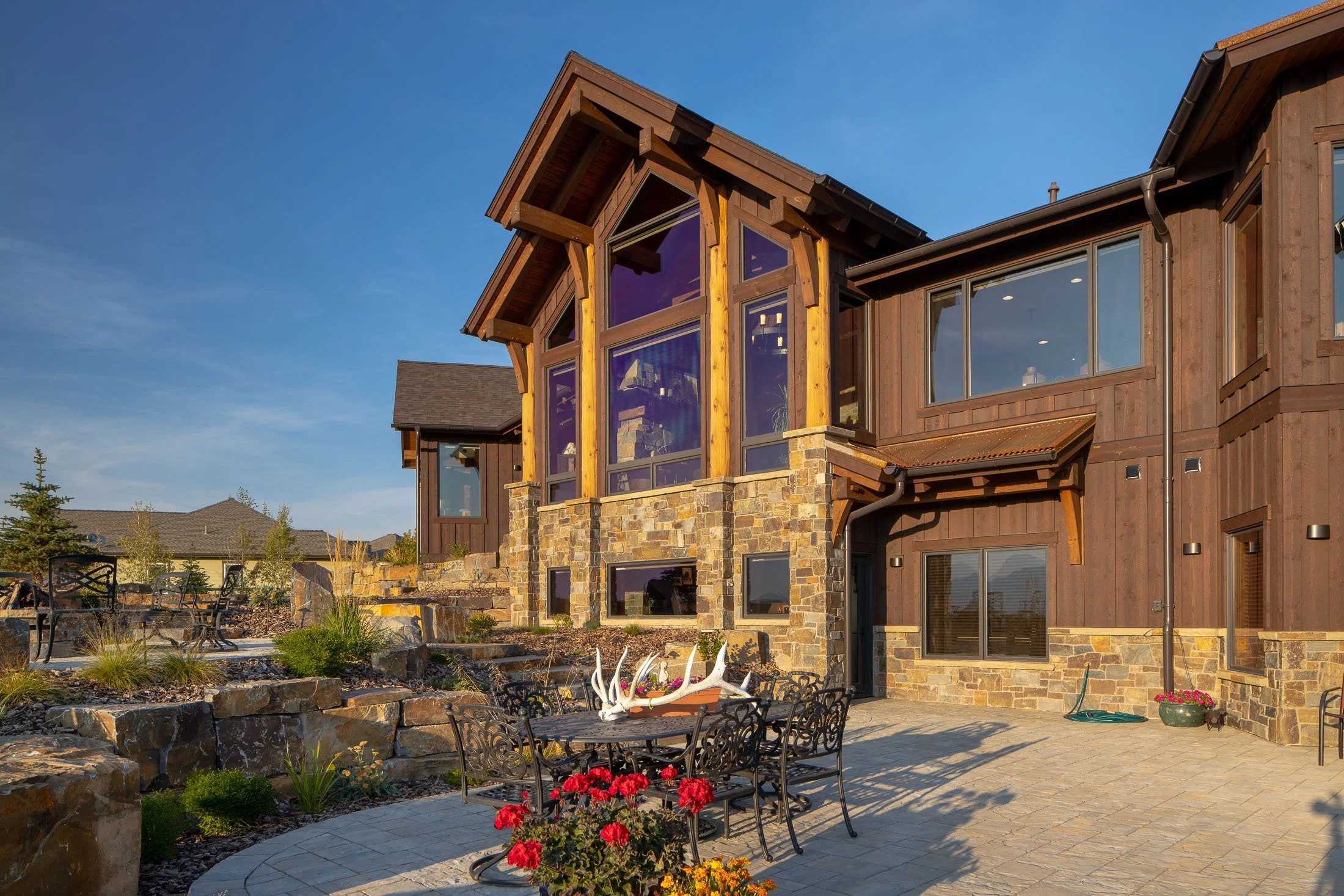 A modern house with wooden and stone exterior, large windows, and a well-maintained patio with outdoor furniture and plants. Custom Home built in Montana by Edgell Building.