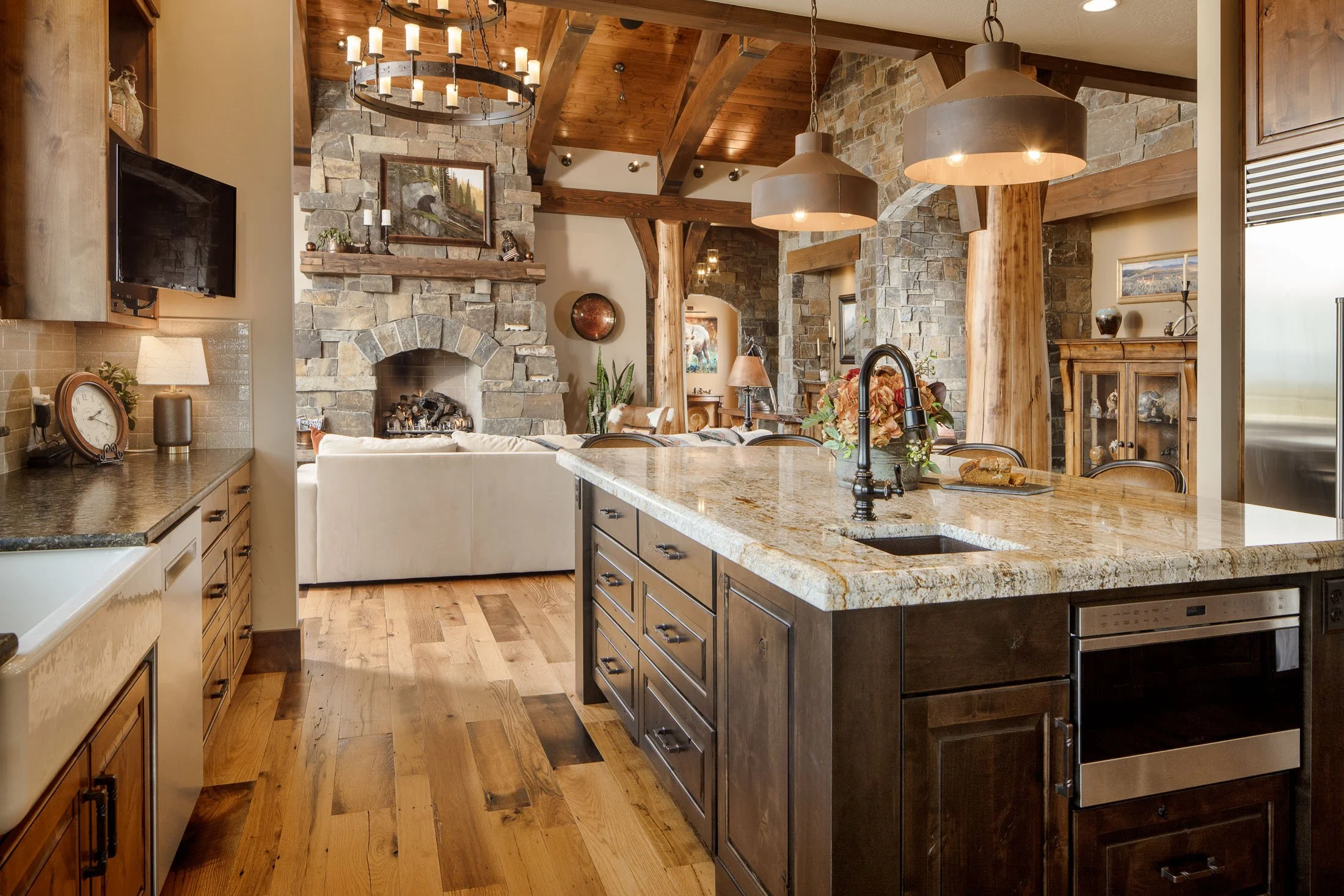 Open-concept kitchen and living area with stone fireplace, wooden beams, granite countertops, and modern appliances. Custom Home built in Montana by Edgell Building.
