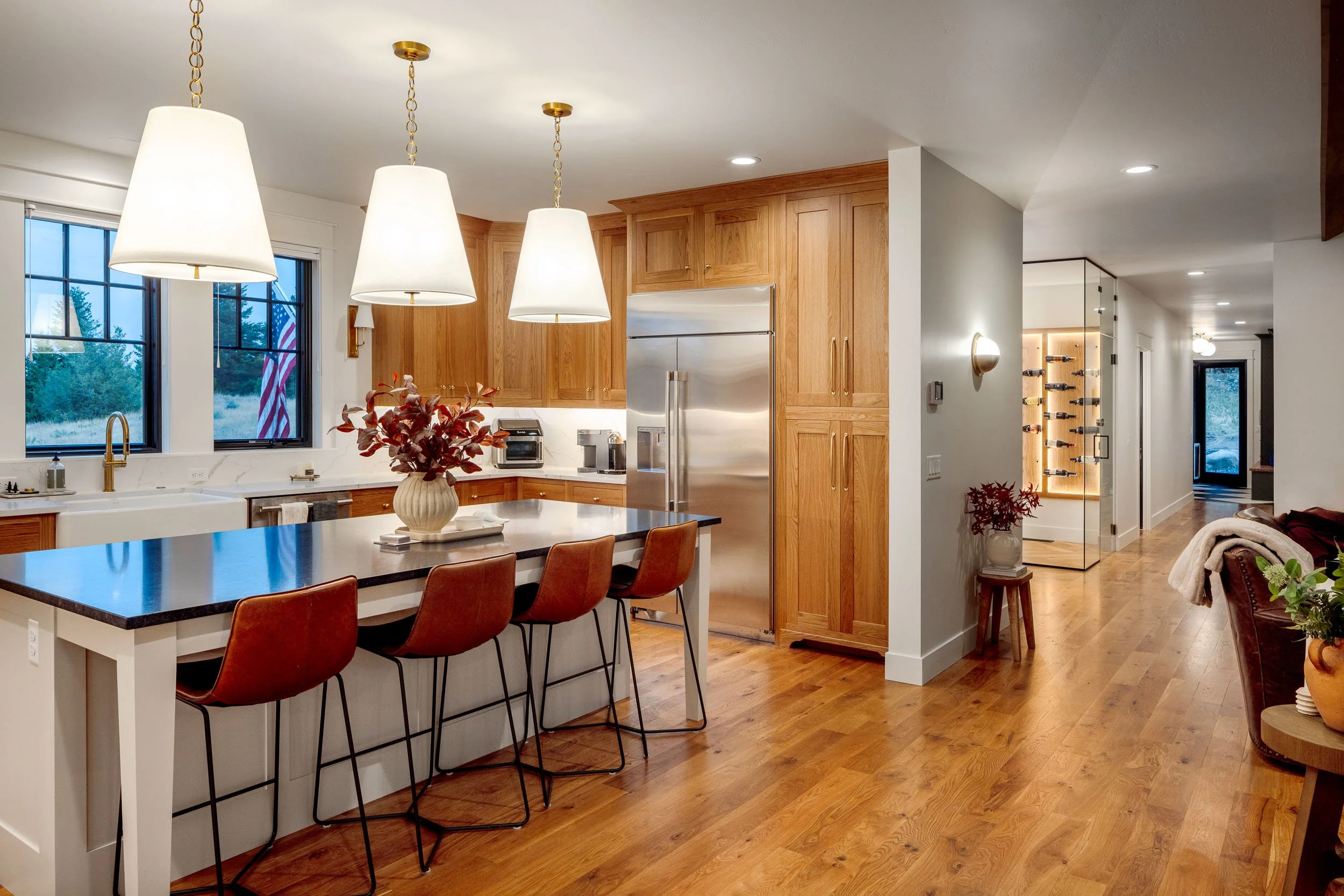 Modern kitchen with wooden cabinets, a white island with a black countertop, orange bar stools, pendant lights, and a view of a hallway with hardwood floors and a wine display.