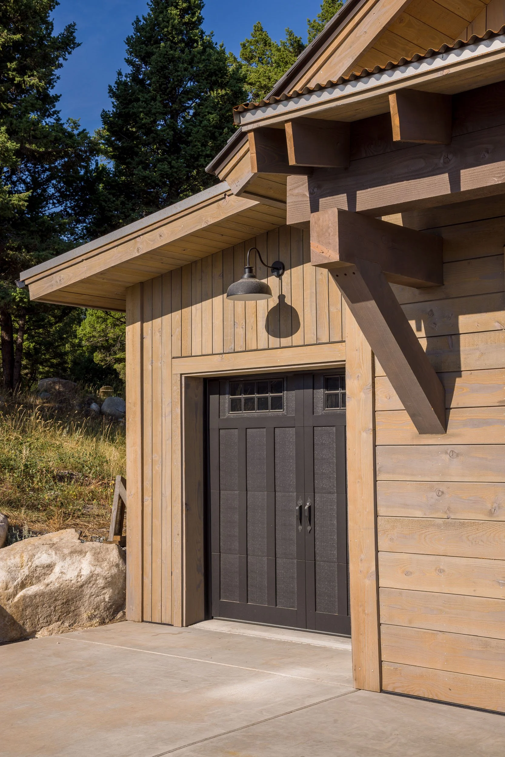 Exterior of a wooden building with a black double door, a black outdoor light fixture above, and a sloped roof with exposed beams, surrounded by trees and rocks. Custom Home built in Montana by Edgell Building.