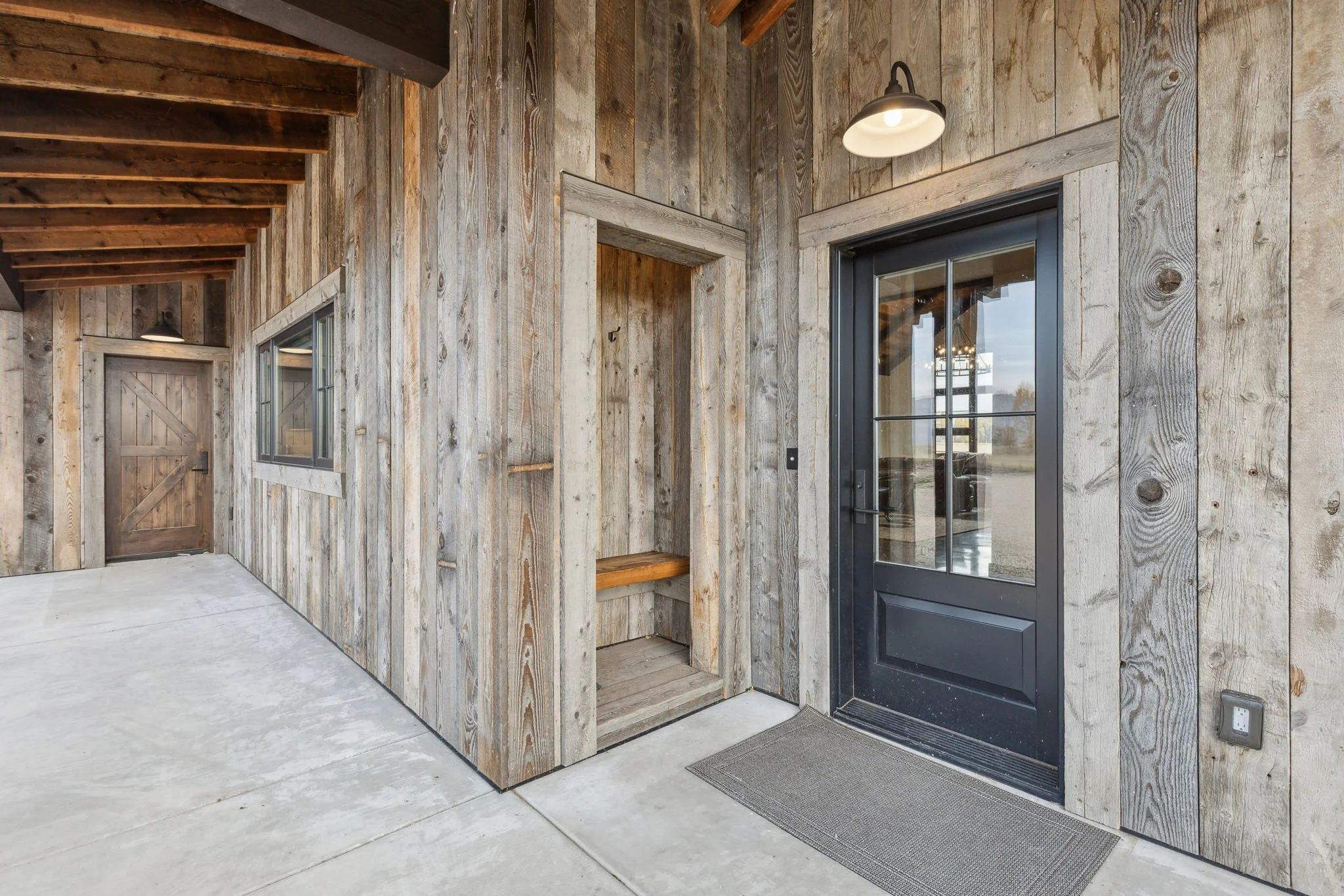 Rustic wooden exterior wall of a building with a black front door, window, and an empty small open closet or storage space with a bench inside. There is a concrete porch and a porch light above. Custom Home built in Montana by Edgell Building.