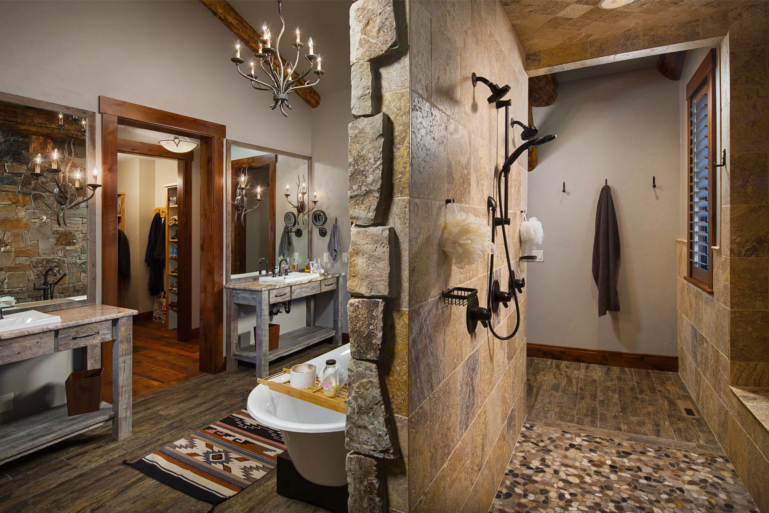 A rustic-style bathroom with a dual vanity, large mirrors, and chandelier lighting on the left side; a private enclosed shower with stone and tile walls, multiple showerheads, and a window. Custom Home built in Montana by Edgell Building.