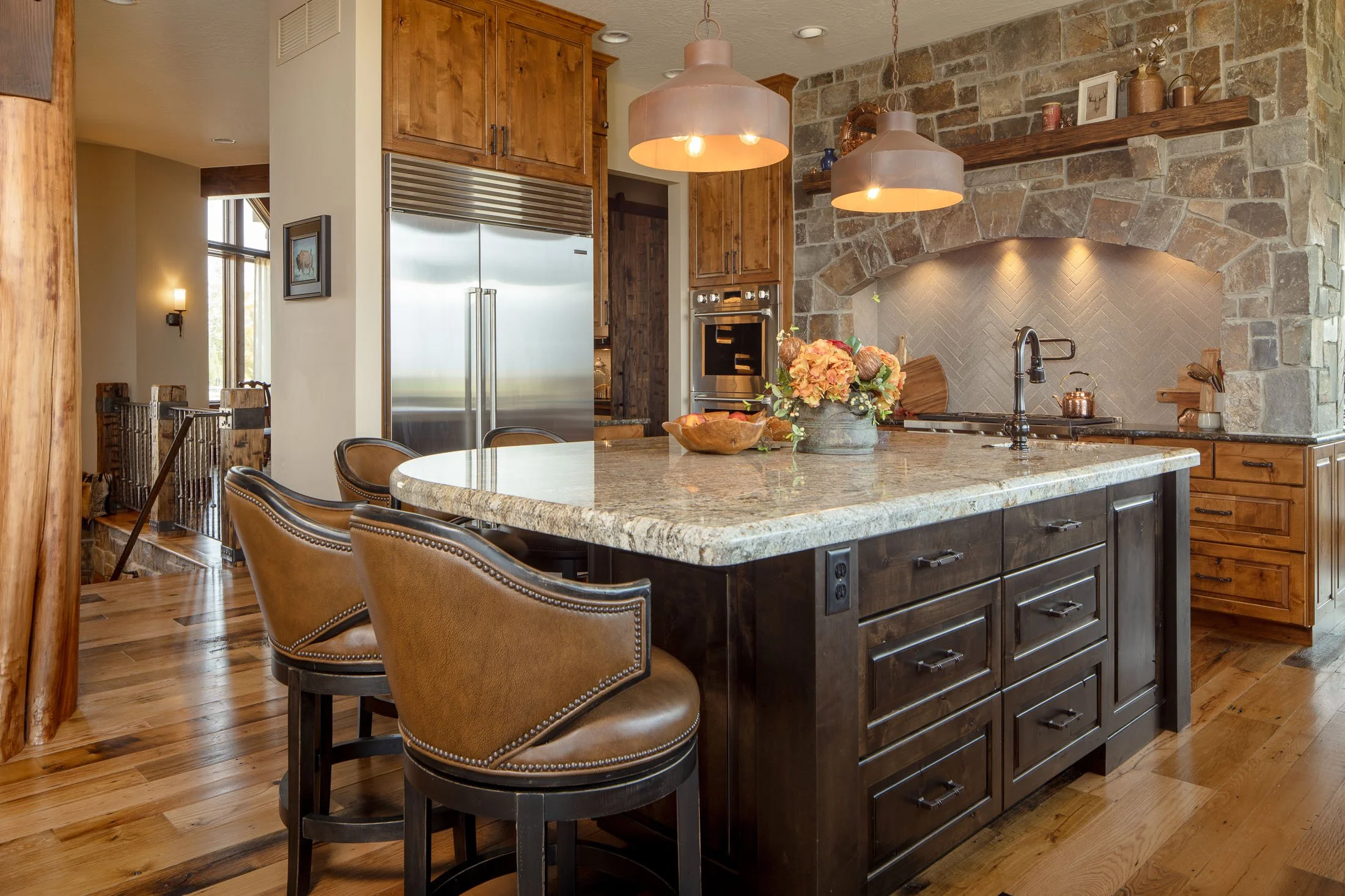Rustic kitchen with granite island, brown leather barstools, wooden cabinets, stainless steel refrigerator, brick wall, and pendant lights. Custom Home built in Montana by Edgell Building.