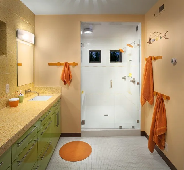 Bathroom with peach-colored walls, green vanity, orange towels on hooks, glass shower with small windows, and decorative fish wall art. Custom Home built in Montana by Edgell Building.