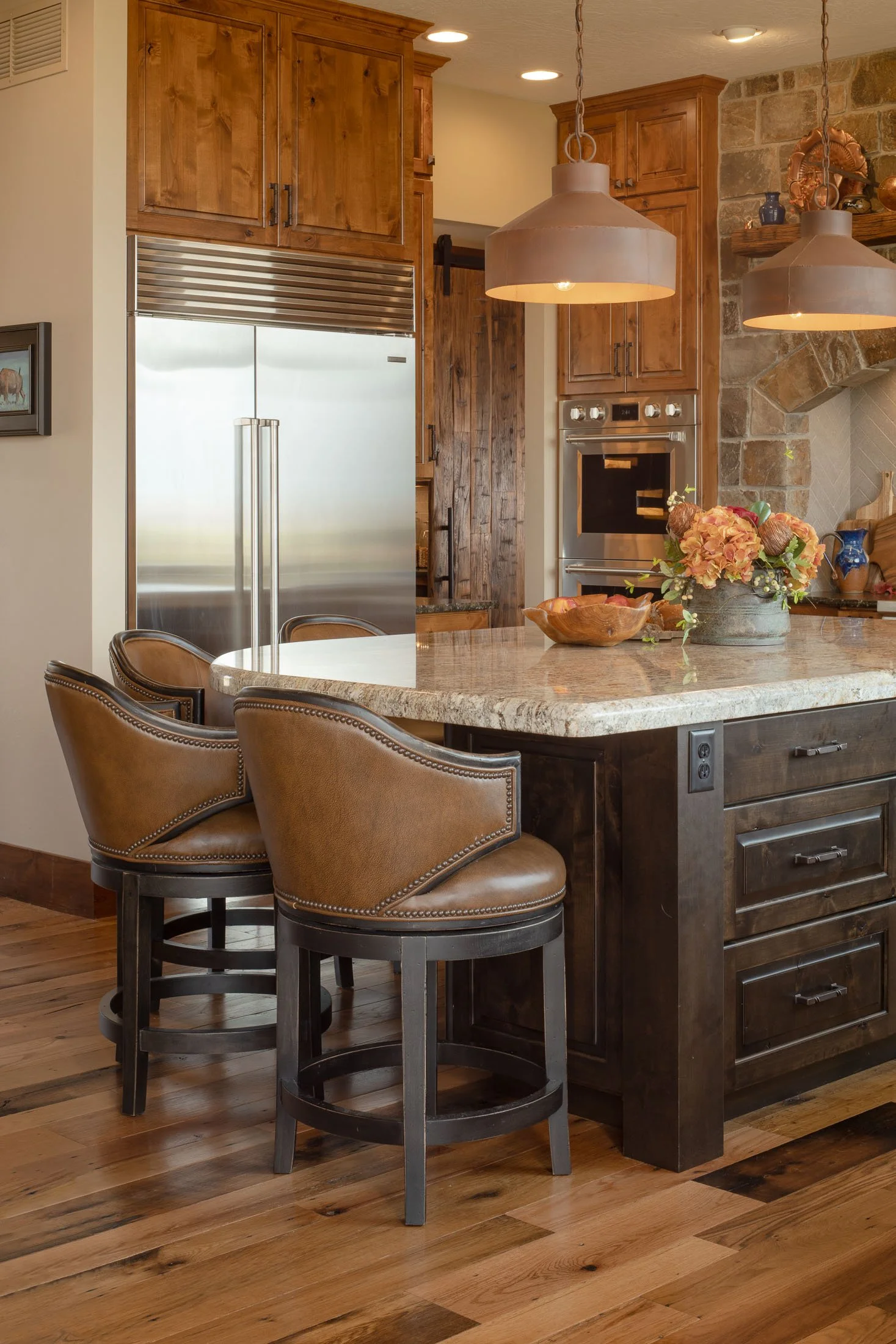 A kitchen featuring a granite island with wooden cabinets, a stainless steel refrigerator, two pendant lights, and a floral centerpiece on the island. Custom Home built in Montana by Edgell Building.