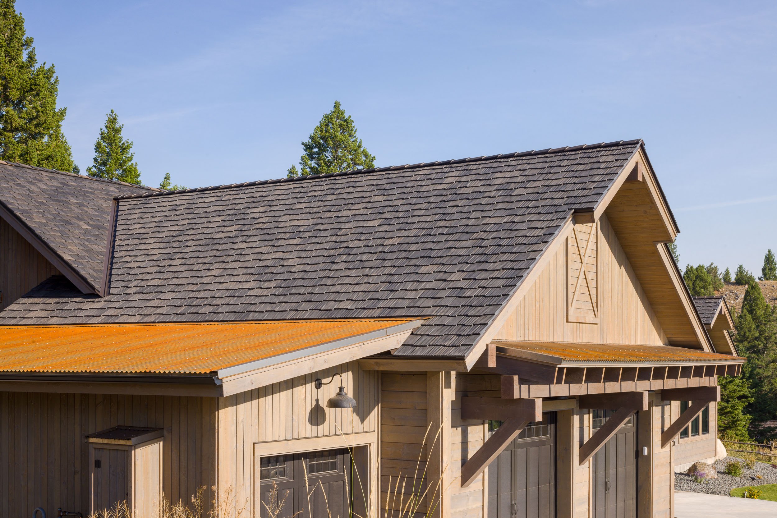 A modern house with a large sloped shingle roof, wooden exterior walls, overhangs supported by wooden brackets, and a garage door visible at the front, surrounded by trees and clear blue sky. Custom Home built in Montana by Edgell Building.