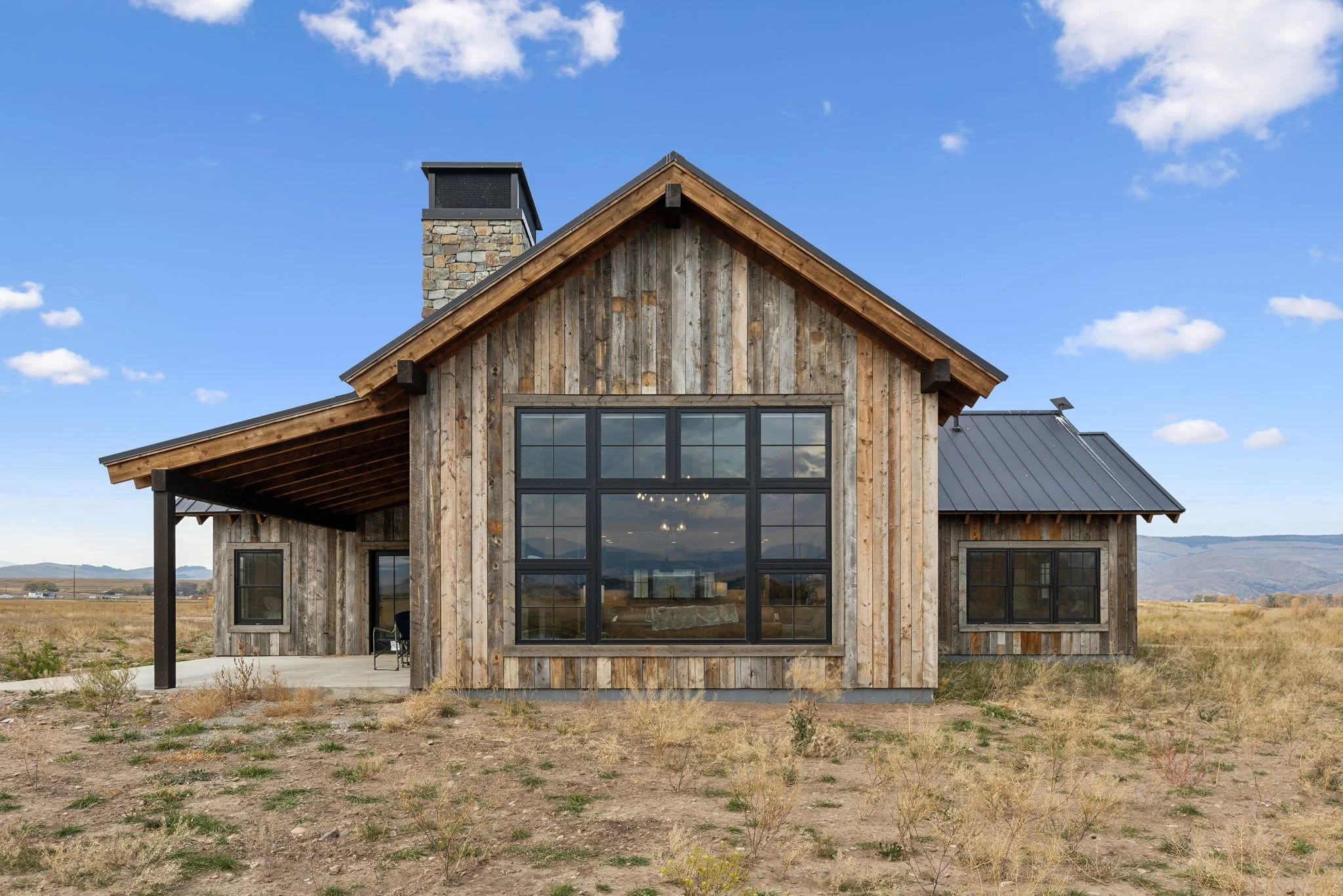 A rustic wooden house with large black-framed windows, a metal roof, and a stone chimney, situated in an open field with mountains in the background under a partly cloudy sky. Custom Home built in Montana by Edgell Building.