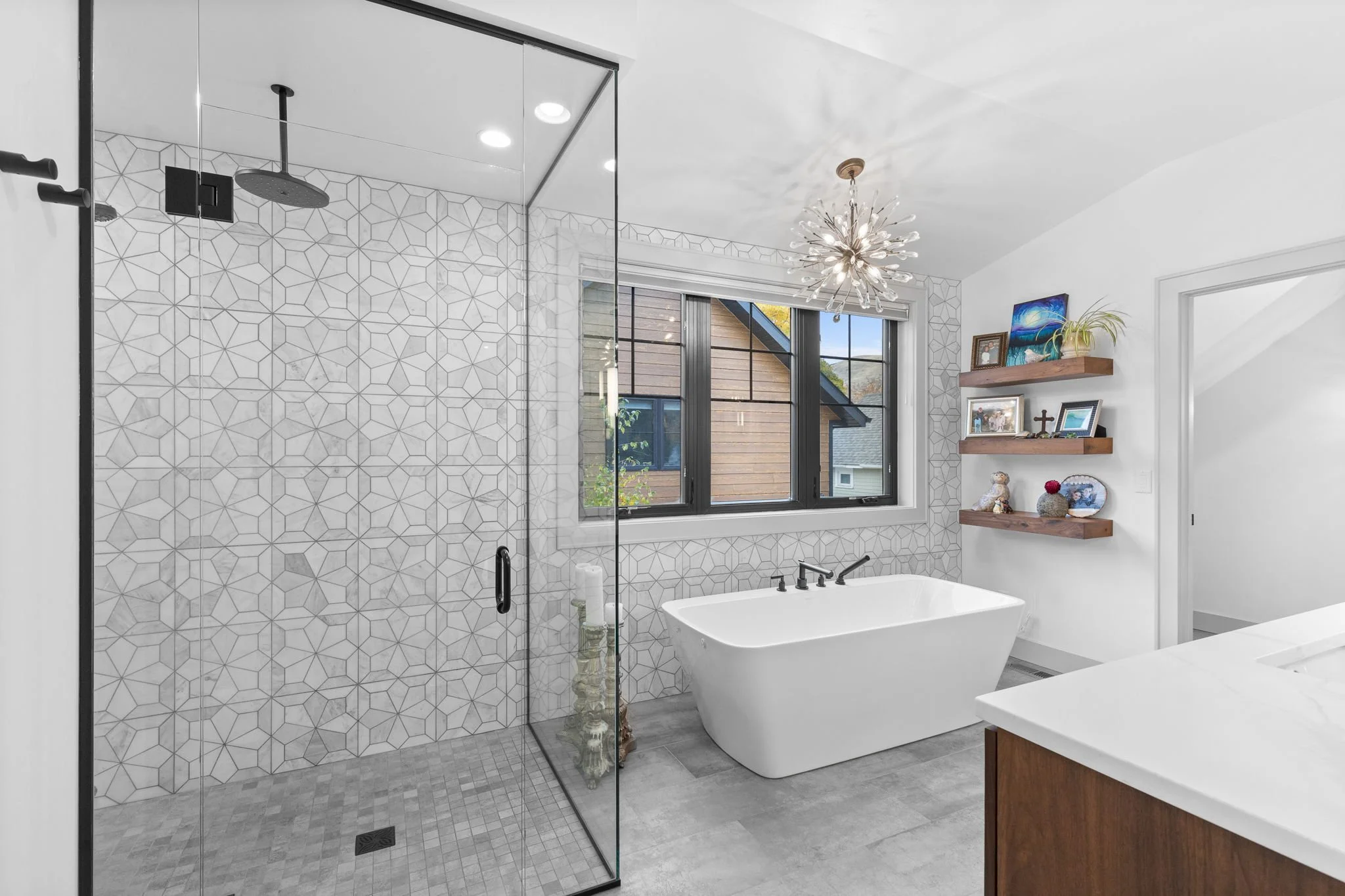 Modern bathroom with a walk-in shower, a freestanding bathtub, patterned tile wall, wooden shelves with decor, a window, and a chandelier. Custom Home built in Montana by Edgell Building.