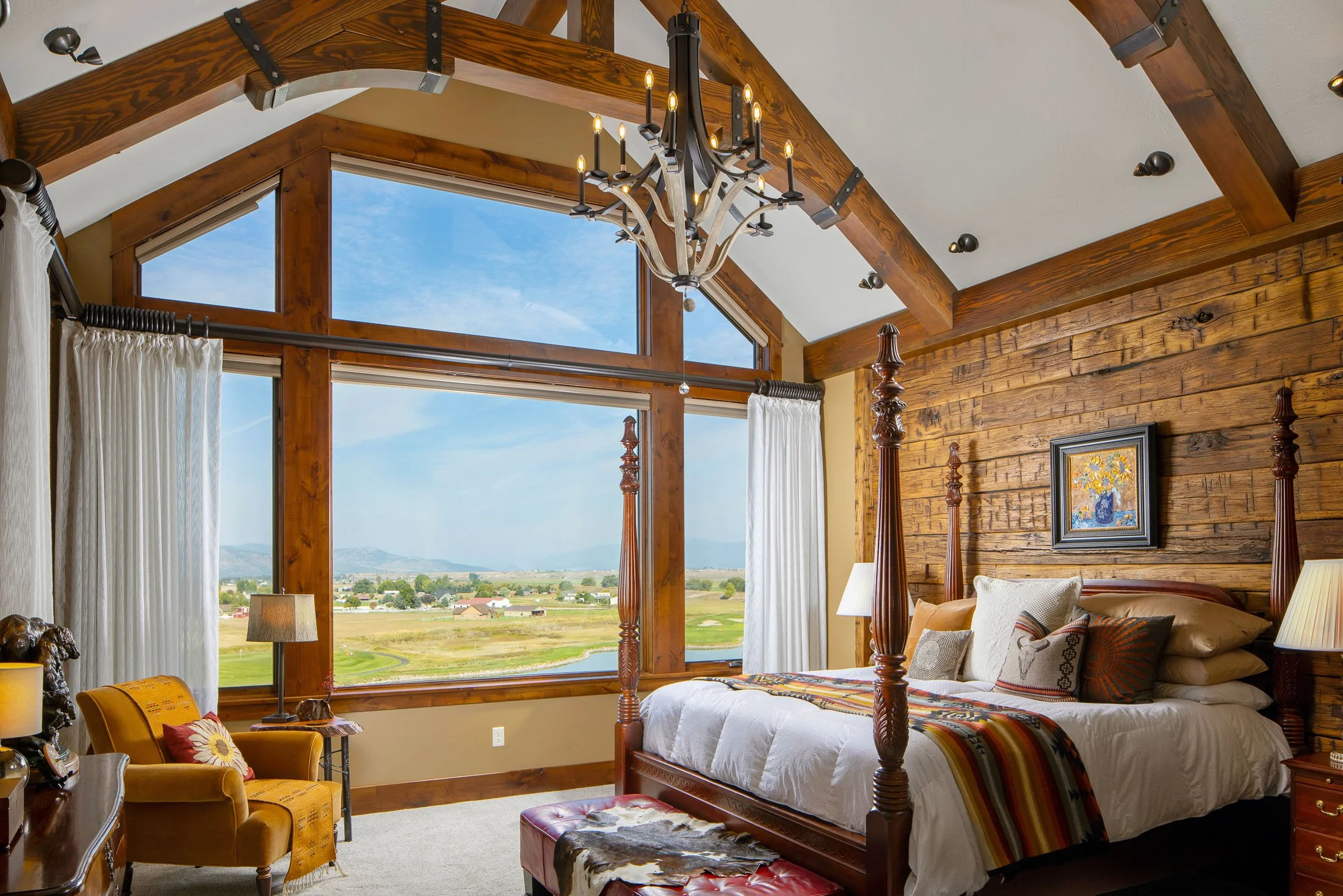 A bedroom with large wooden framed windows showing a scenic view of fields and mountains, a wooden four-poster bed with white bedding and decorative pillows, and various lamps and furniture. Custom Home built in Montana by Edgell Building.