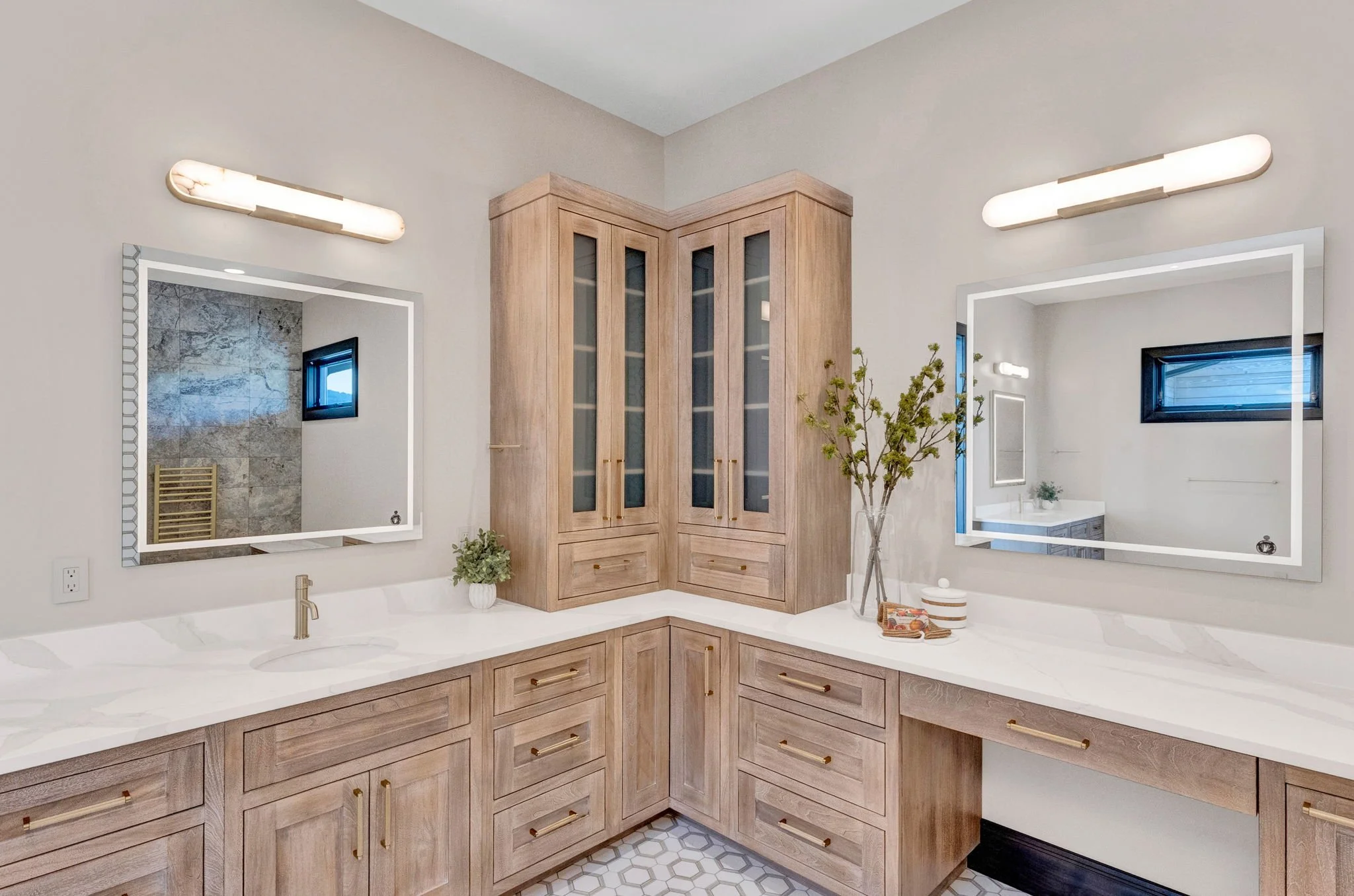 A modern bathroom with a corner vanity featuring a white countertop, two mirrors with white frames, and wooden cabinetry. There are two light fixtures above each mirror, a small window on each side. Custom Home built in Montana by Edgell Building.