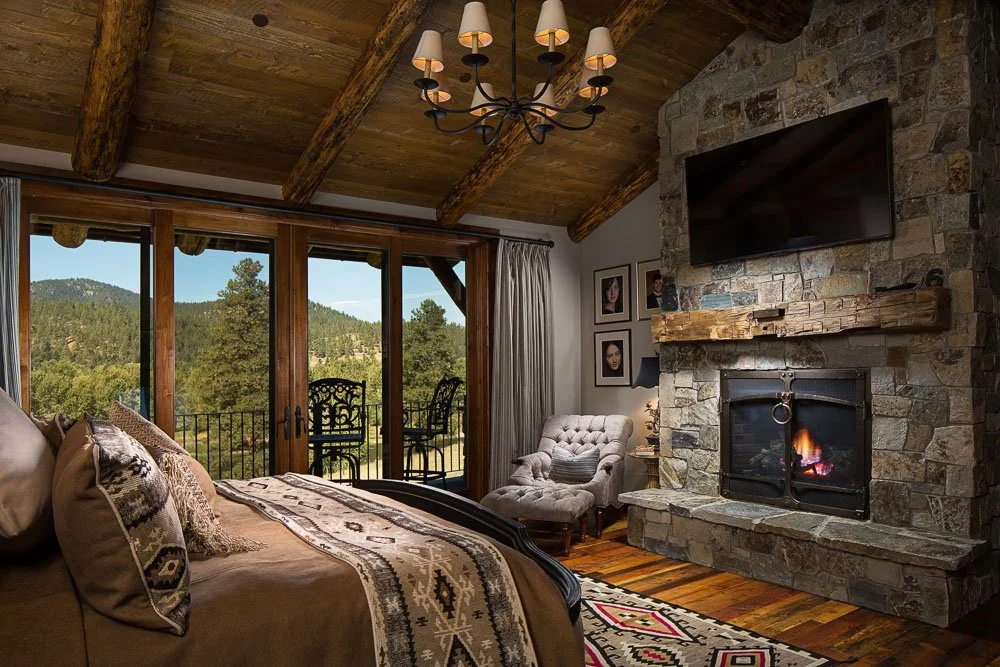 Cozy bedroom with a large bed decorated with patterned pillows, a stone fireplace with a fire, a wall-mounted TV above the fireplace, a tufted armchair, a wooden side table, framed photos on the wall, large glass doors leading to a balcony with outdo