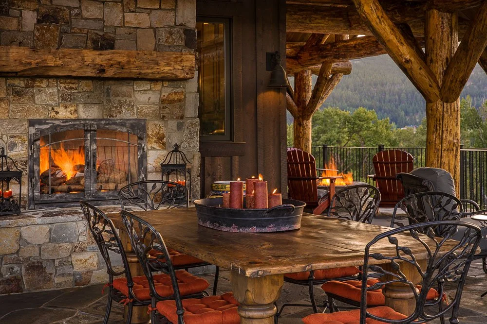 Outdoor patio area with a stone fireplace, wooden dining table with metal chairs, and a scenic view of trees and mountains. Custom Home built in Montana by Edgell Building.