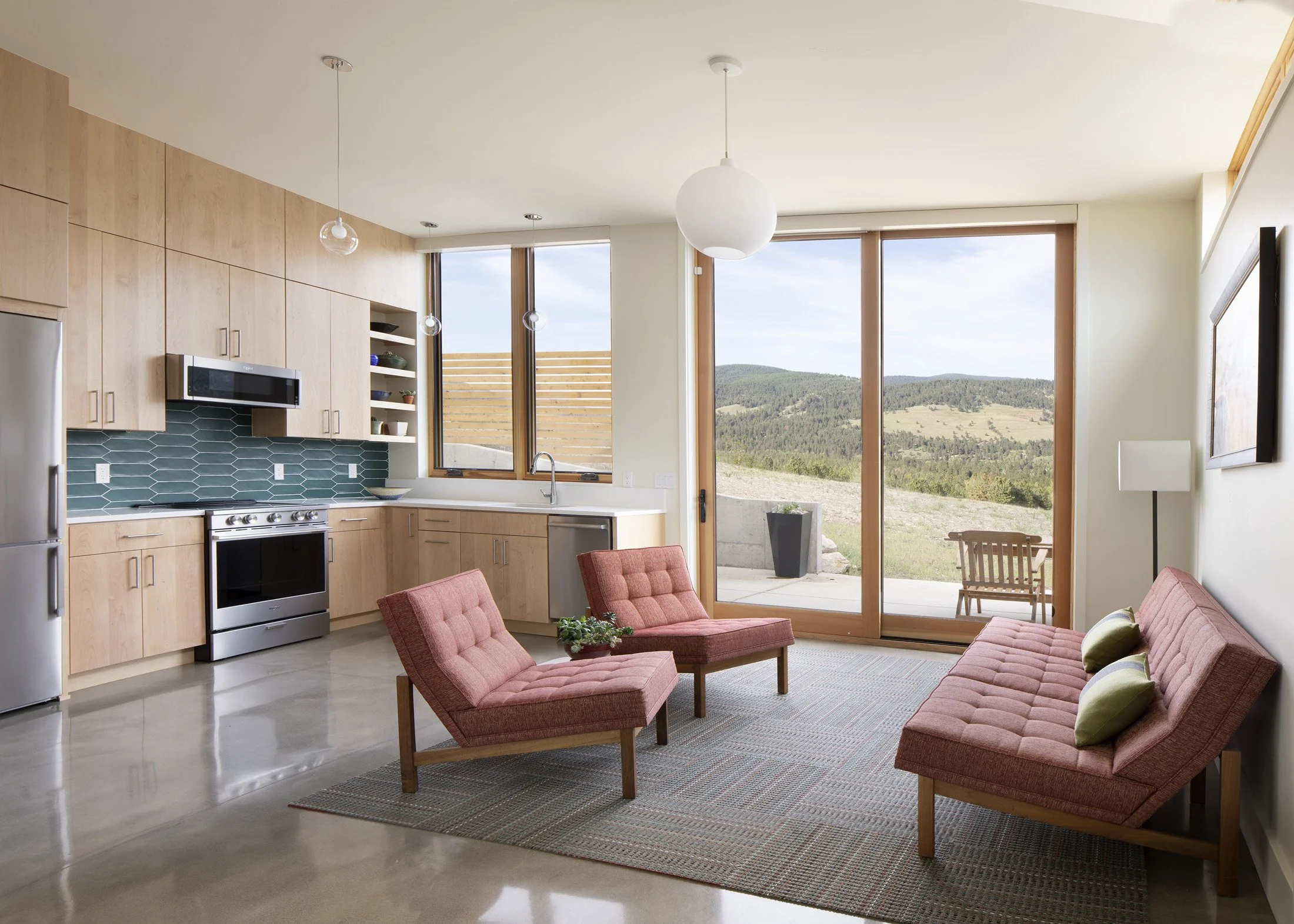 Living room with pink mid-century modern sofa and chairs, wooden kitchen cabinets, green tiled backsplash, large sliding glass door to outdoor patio, mountain view. Custom Home built in Montana by Edgell Building.