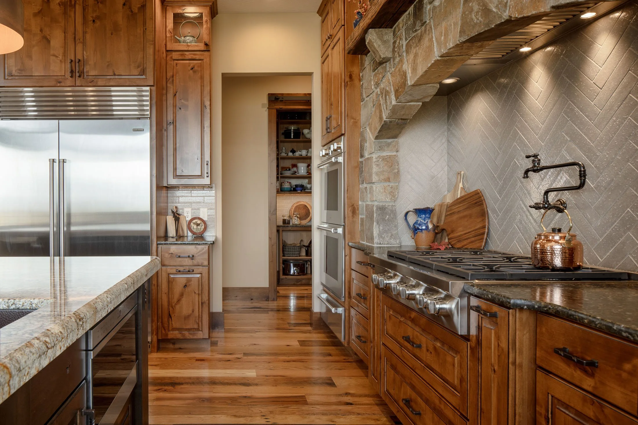 A cozy kitchen with wooden cabinets, a stainless steel refrigerator, a stove with copper kettle, and a stone arch over the stove. Open shelves with dishes and a small pantry in the background. Custom Home built in Montana by Edgell Building.