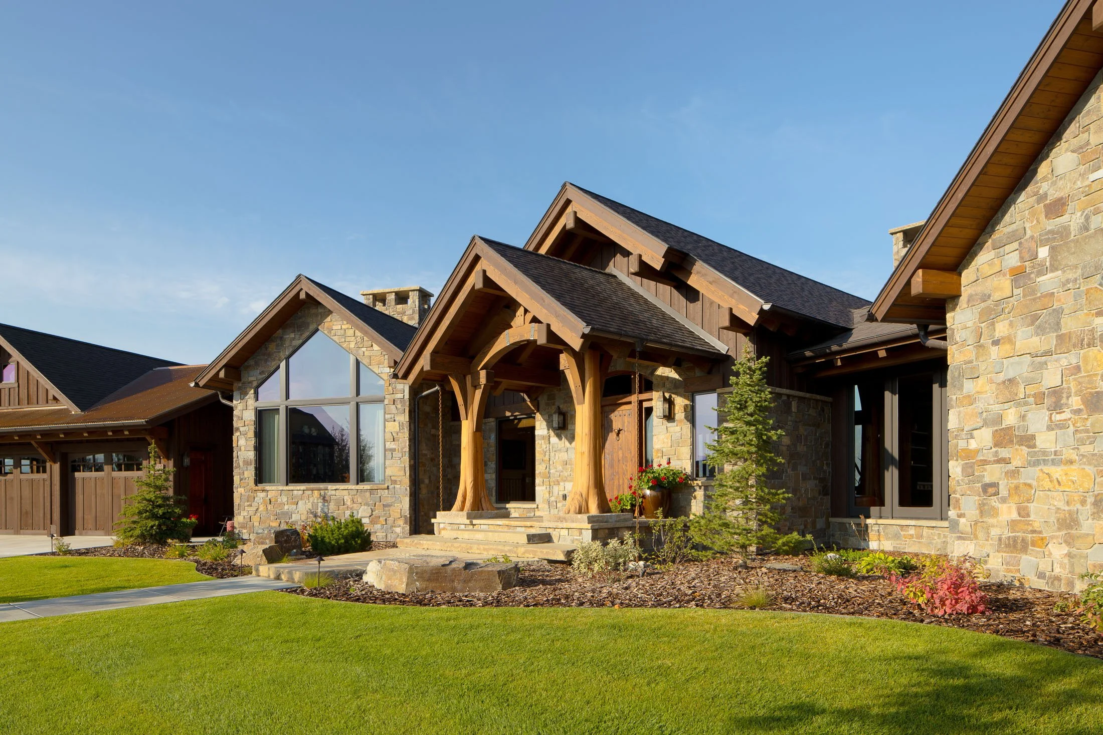 A modern house with a stone and wood exterior, large windows, a front porch with wooden beams, and a manicured lawn with small trees and bushes under a clear blue sky. Custom Home built in Montana by Edgell Building.