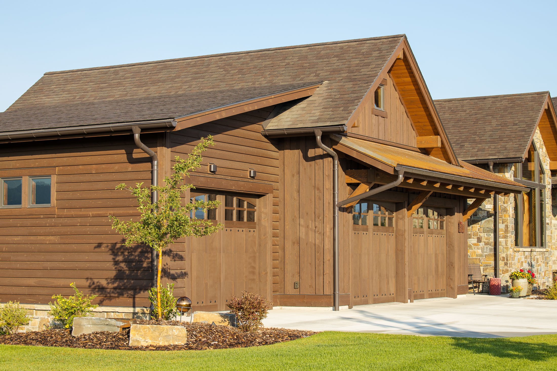 A residential house featuring a wooden exterior, large garage doors, a sloped roof with shingles, and a landscaped front yard with garden plants and a small tree. Custom Home built in Montana by Edgell Building.