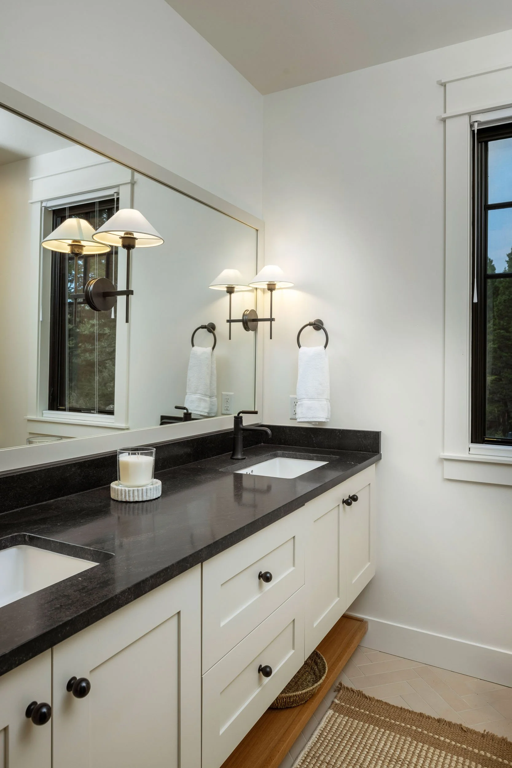 A bathroom vanity with a black countertop, white cabinets, and black knobs. A candle sits on the counter. A large mirror is mounted above. Two wall-mounted light fixtures with multiple bulbs and white shades flank the mirror. A towel hangs on a black