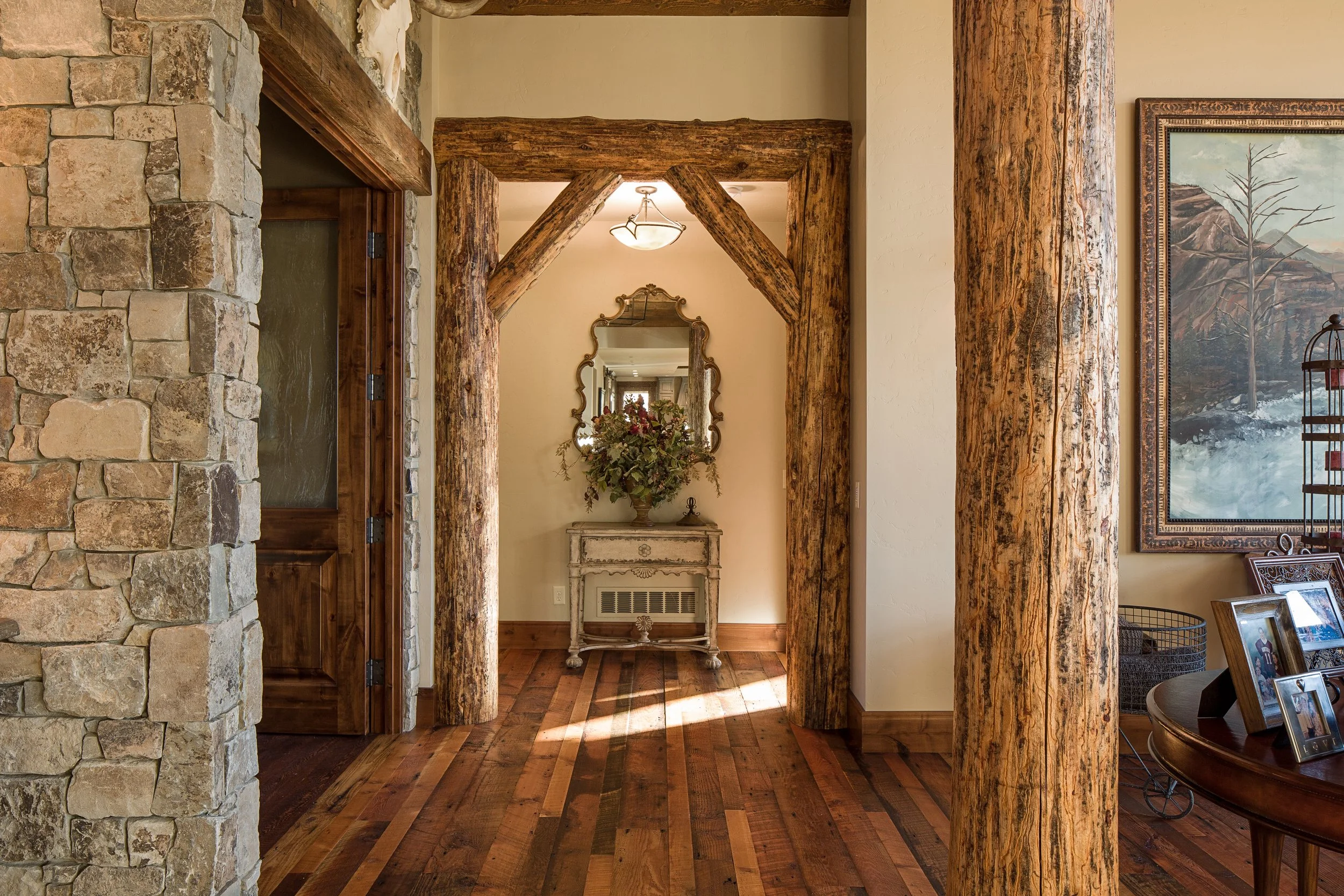 Tile and wood floor hallway with stone wall on left, rustic wooden beams framing doorway, decorative mirror, table with flowers, and framed landscape painting. Custom Home built in Montana by Edgell Building.
