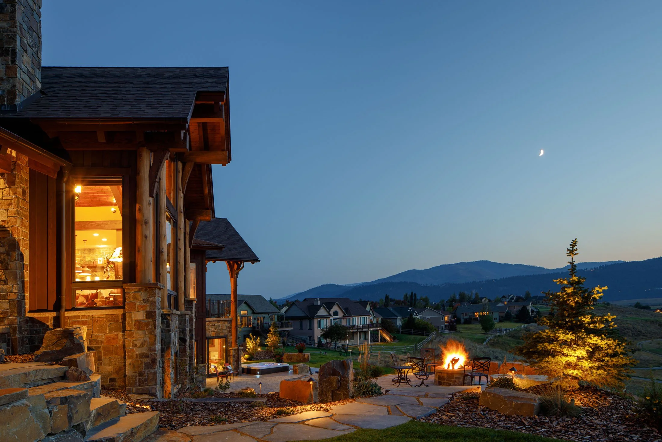 View of a mountain landscape at dusk with a house showing lit windows and a fire pit in the yard, a young pine tree illuminated nearby, and a crescent moon in the sky. Custom Home built in Montana by Edgell Building.