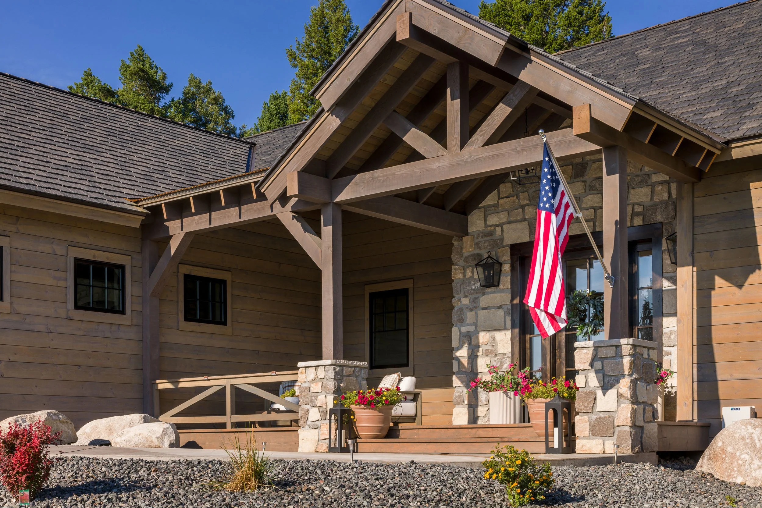 Front porch of a house with wooden and stone exterior, American flag hanging from a pole, porch furniture, and potted flowers, under a clear blue sky. Custom Home built in Montana by Edgell Building.