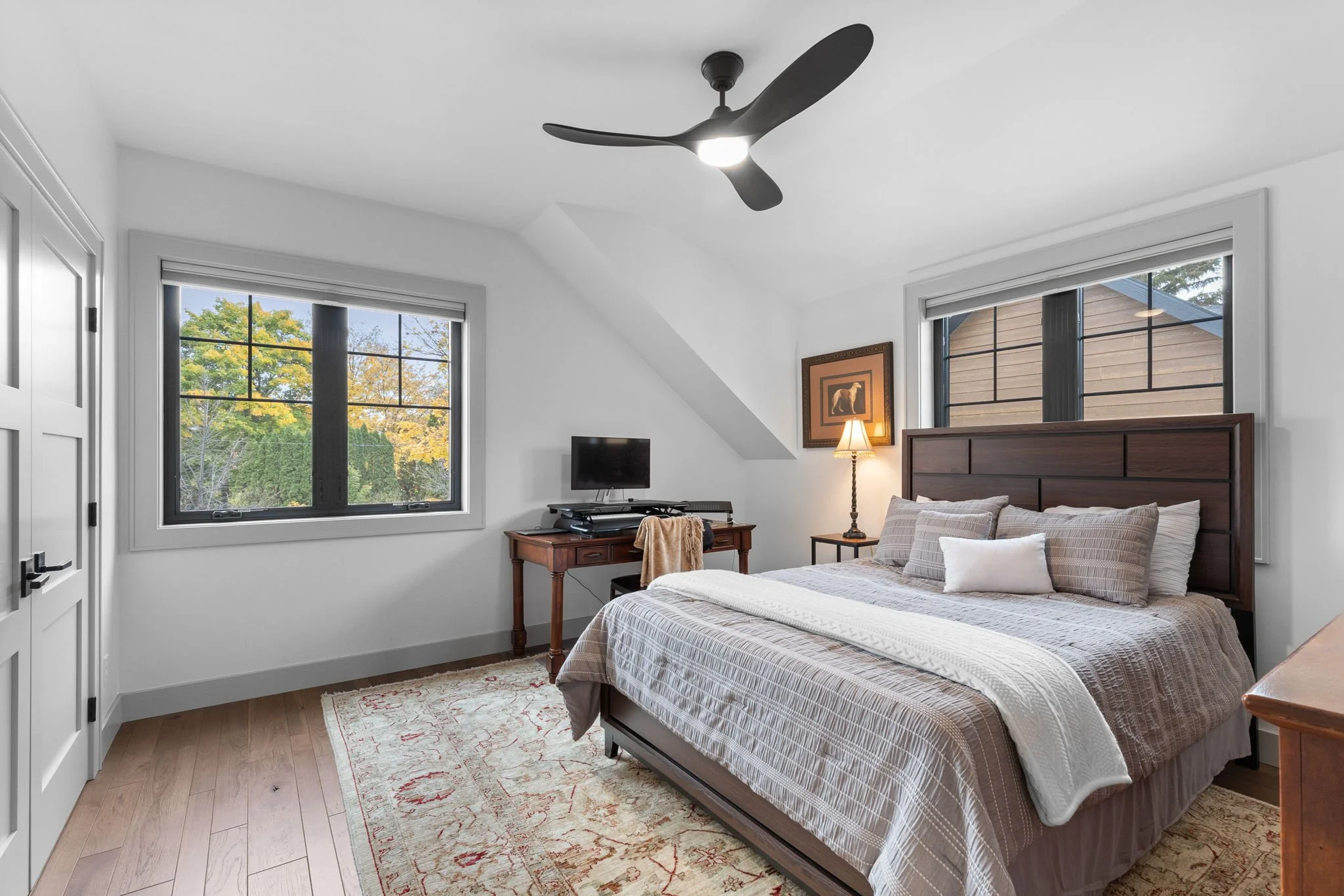 Bedroom with white walls, hardwood floor, large bed with beige bedding and pillows, dark wooden headboard, side table with lamp, window showing trees, ceiling fan. Custom Home built in Montana by Edgell Building.