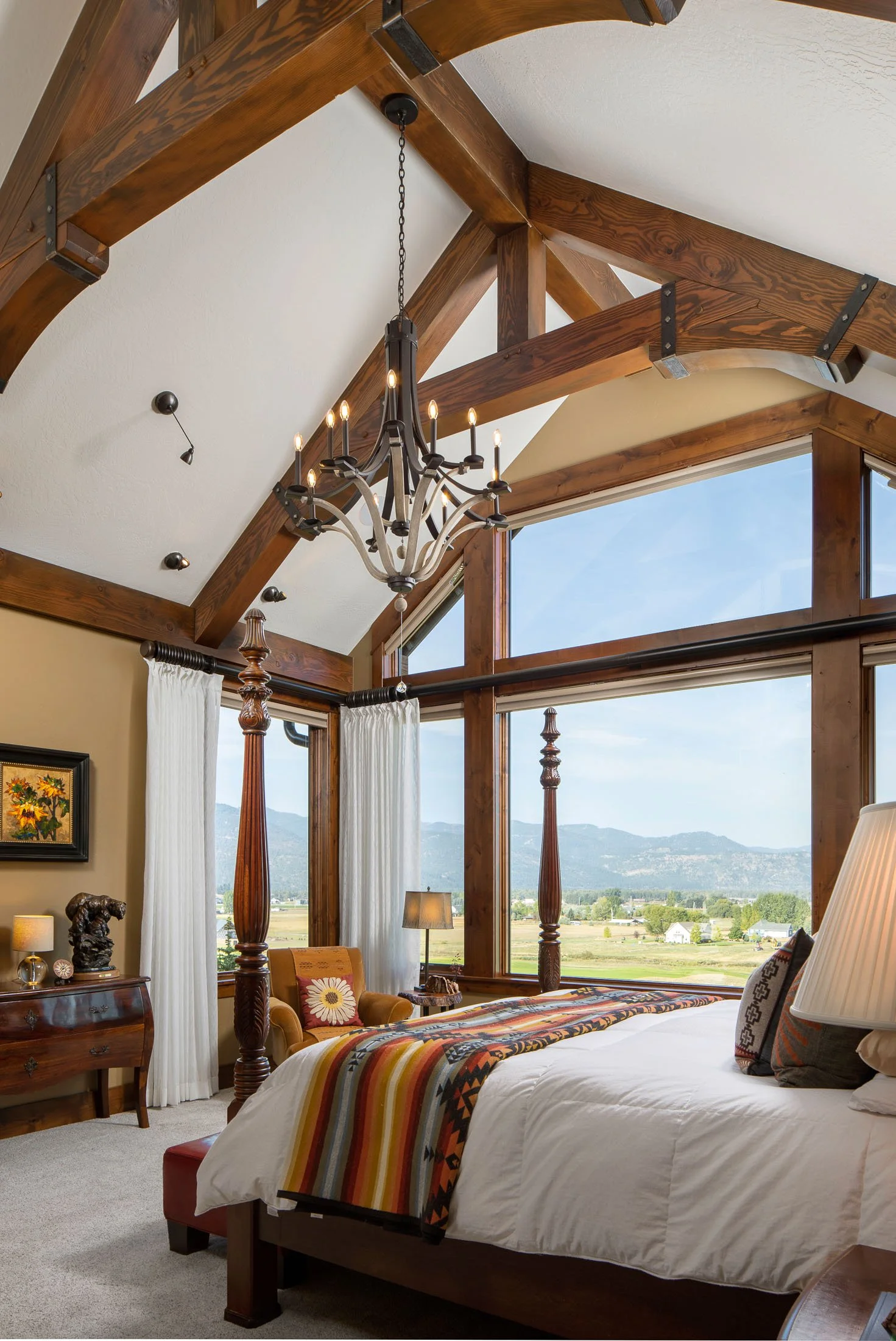 A bedroom with large windows showing mountains and fields outside, featuring a wood beam ceiling, a chandelier, a bed with a colorful striped blanket, a nightstand with a lamp, and a wooden armchair. Custom Home built in Montana by Edgell Building.