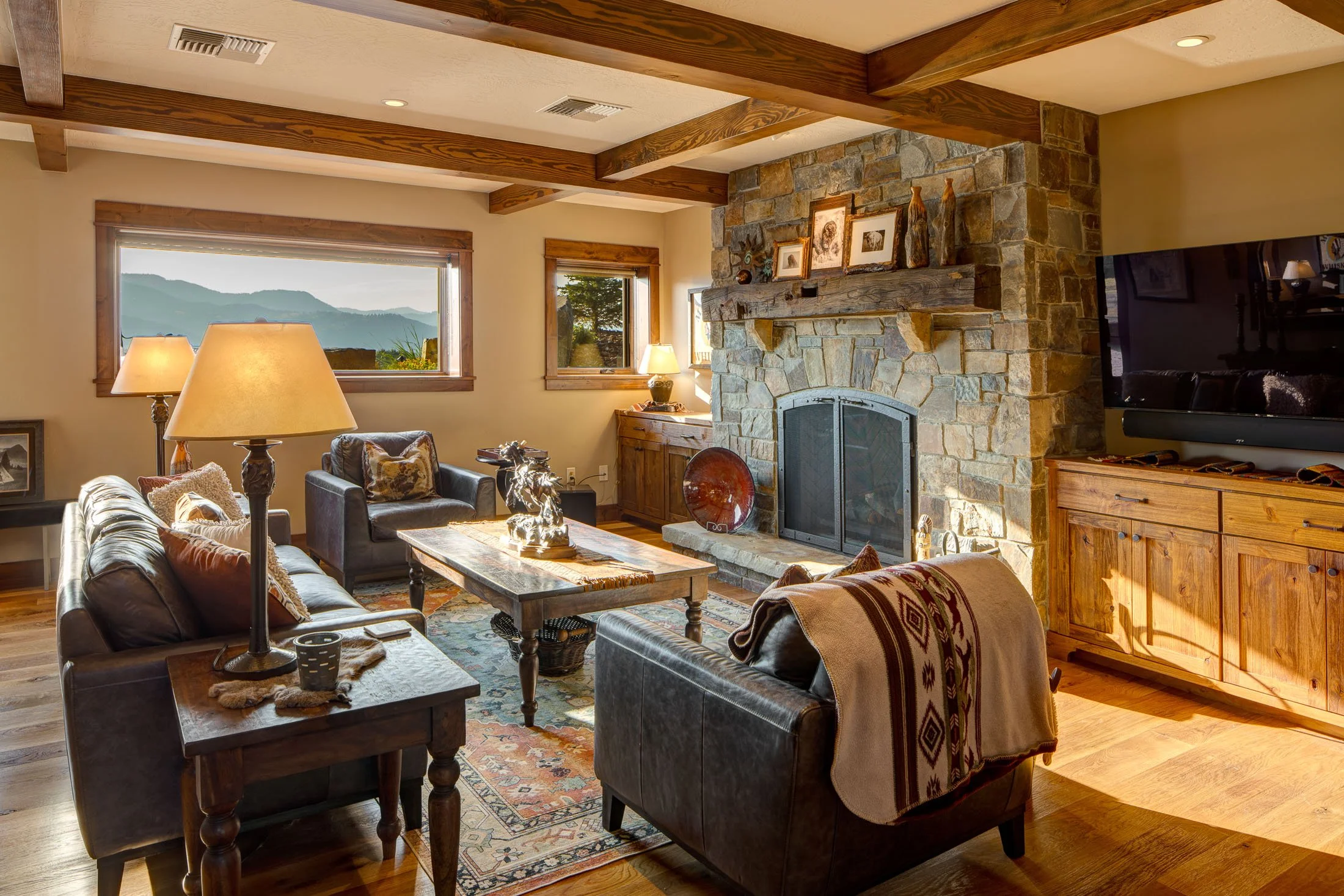 Cozy living room with a stone fireplace, wooden beams, and a view of mountains through the window. It features a leather sofa, wooden coffee table, armchairs, lamps, and a flat-screen TV. Custom Home built in Montana by Edgell Building.